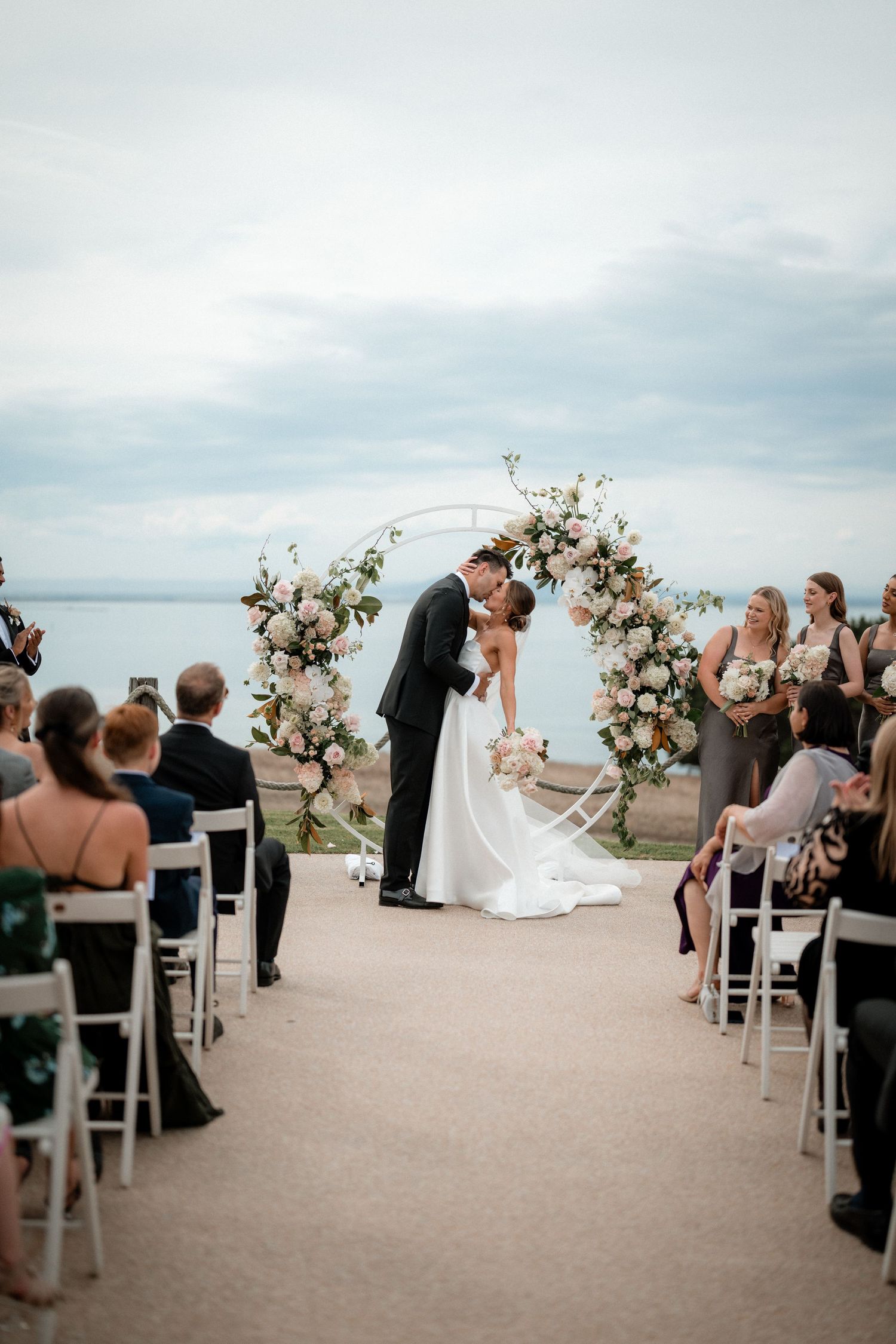 Romantic waterfront wedding ceremony with couple under floral arch and guests seated on white chairs overlooking lake view.