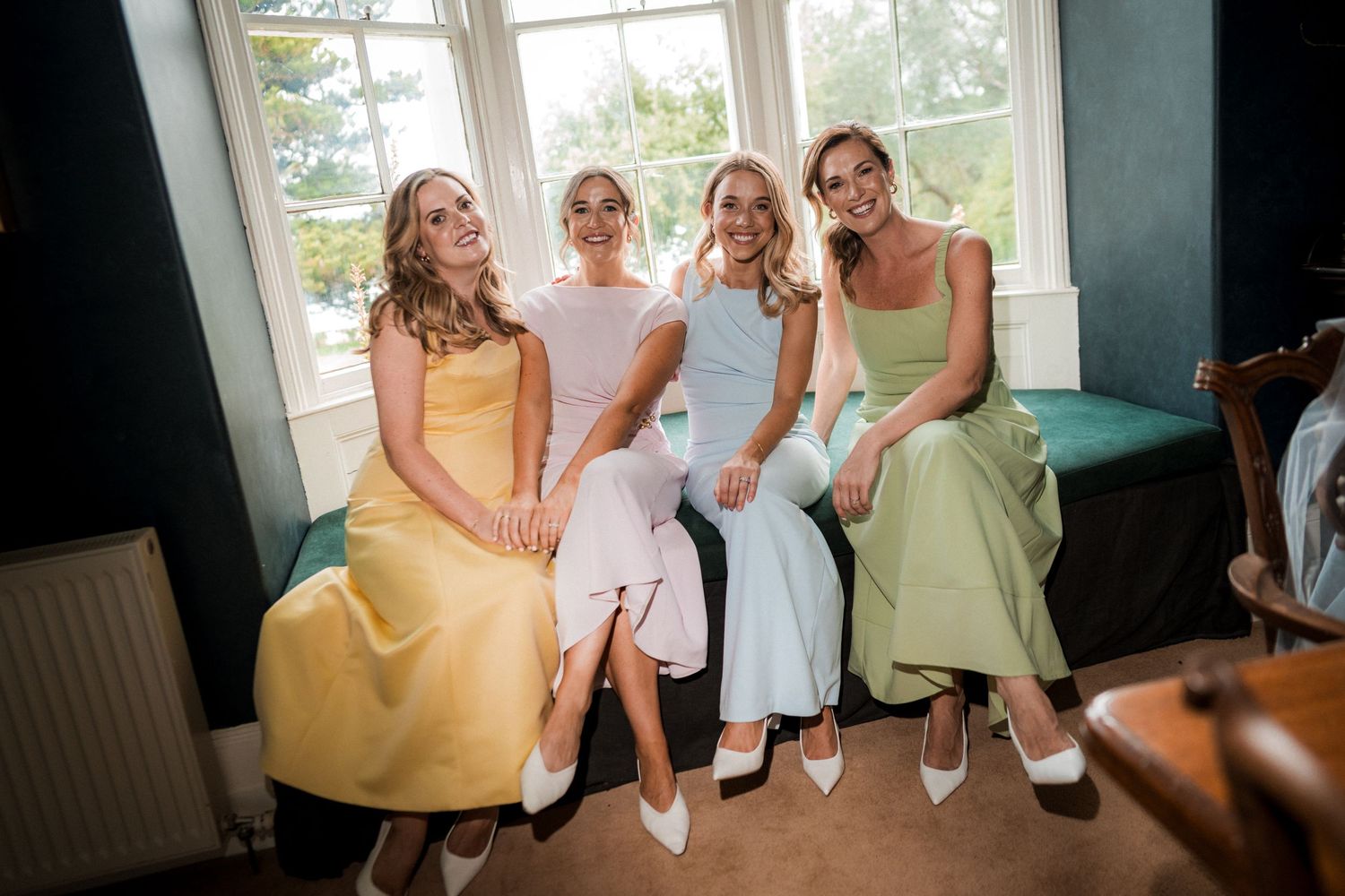 Four people wearing pastel colored dresses in yellow, white, blue and mint green seated together in a bright room.