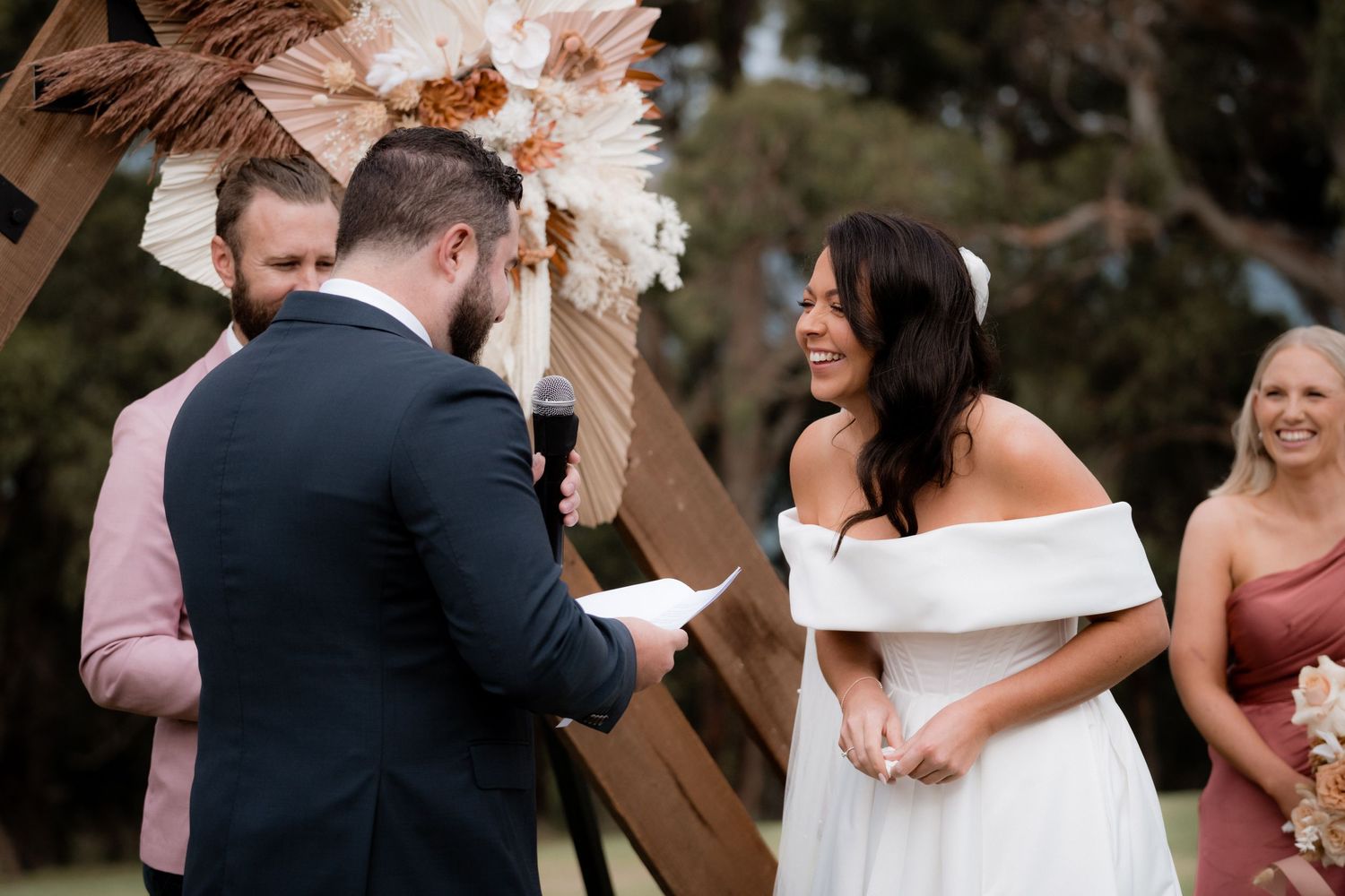 A couple stands at a wooden arch during an outdoor wedding ceremony with a wedding party member in pink looking on.