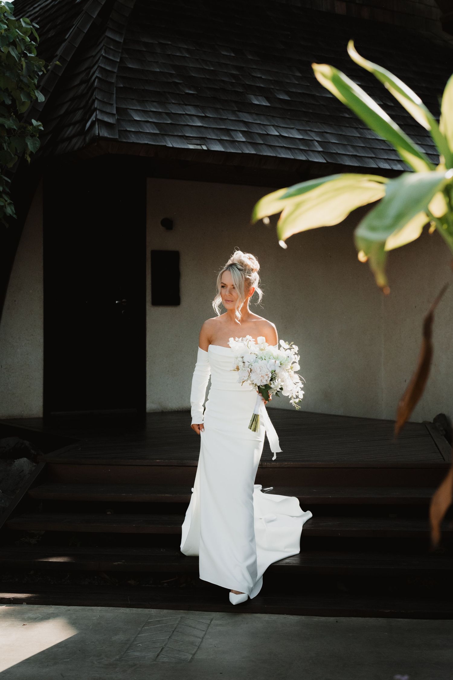 Elegant off-shoulder wedding dress with long sleeves and white bouquet against tropical backdrop.