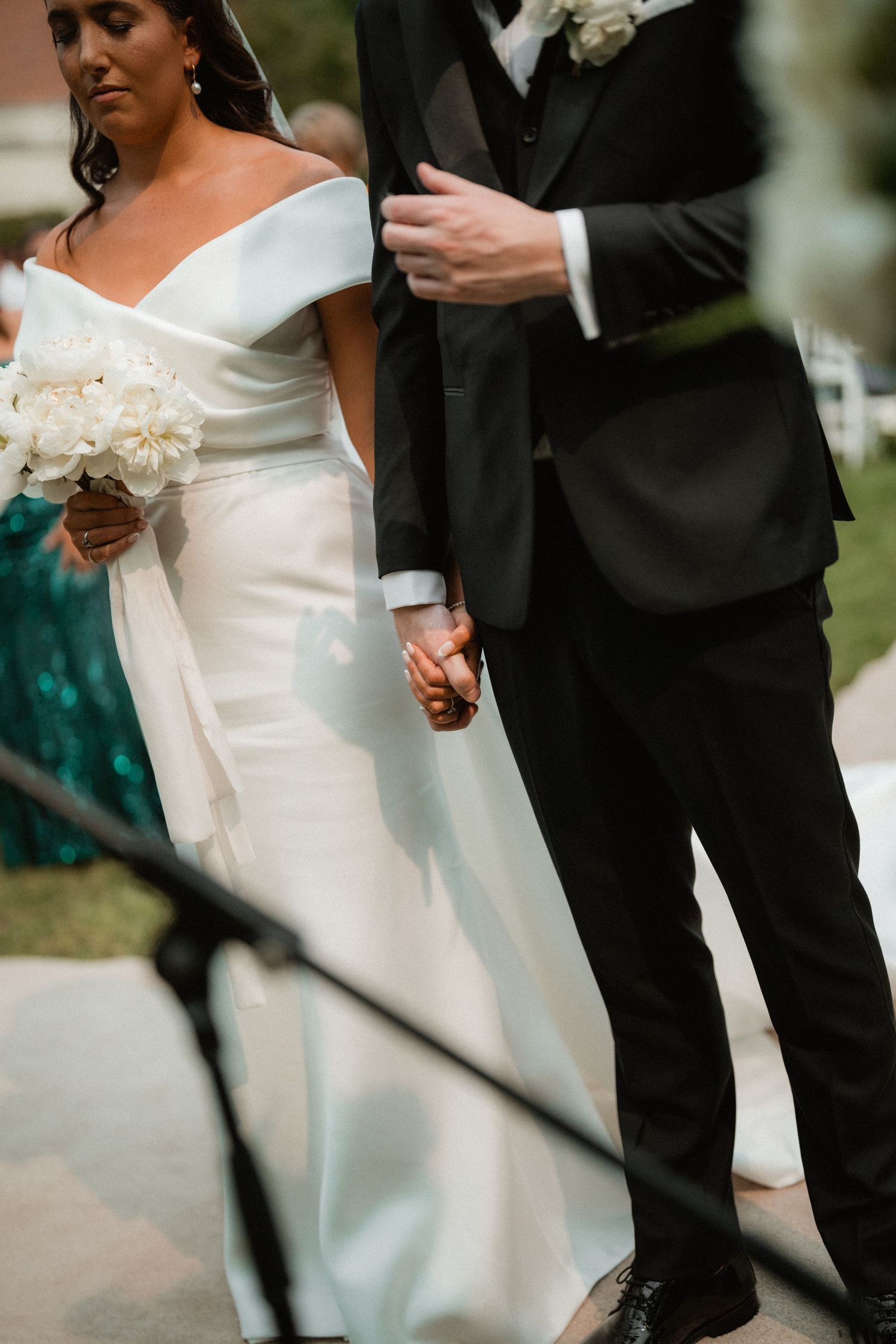 Newlywed couple holding hands at outdoor wedding ceremony, showing elegant white gown and black tuxedo details.
