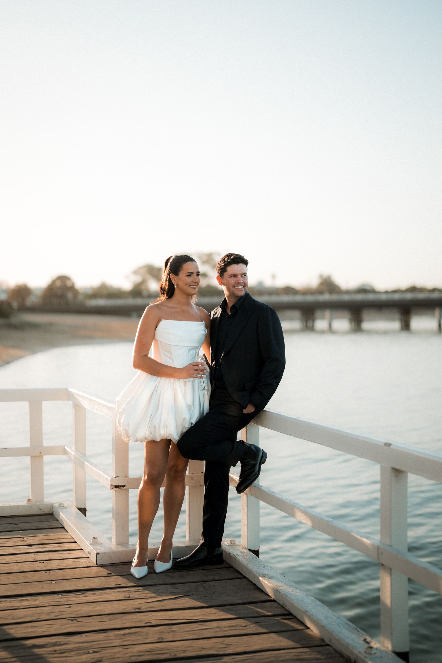 A couple in formal attire poses together on a wooden pier at sunset with a bridge visible in the background.