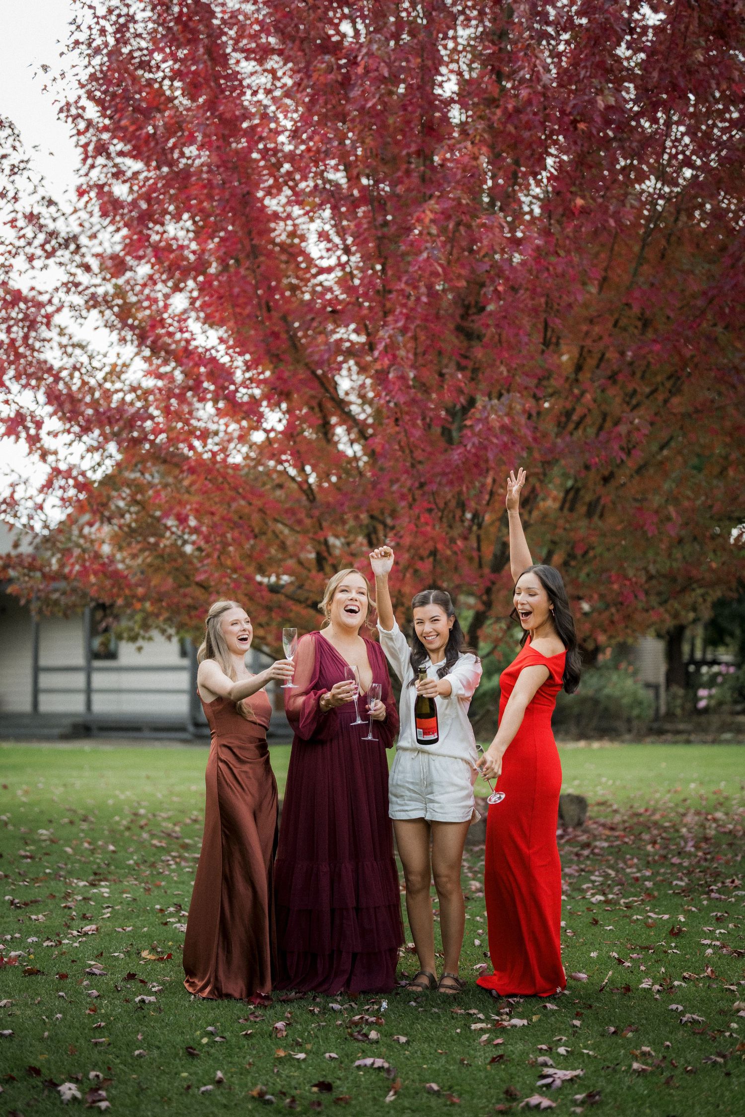 Four bridesmaids in red and burgundy gowns celebrating with champagne under vibrant autumn red foliage.