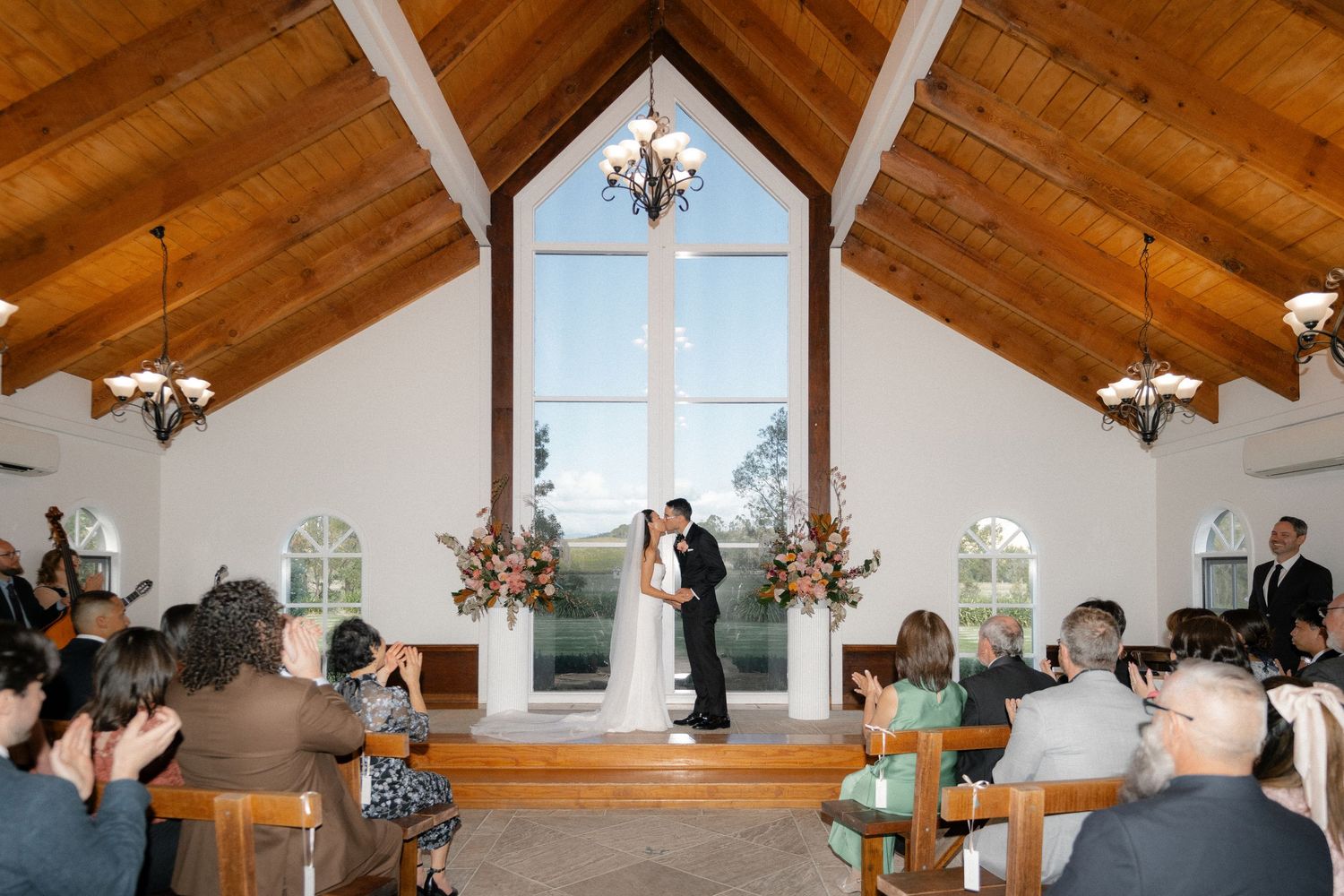 Wedding ceremony in white chapel with vaulted wood ceiling, couple kissing at altar before guests.