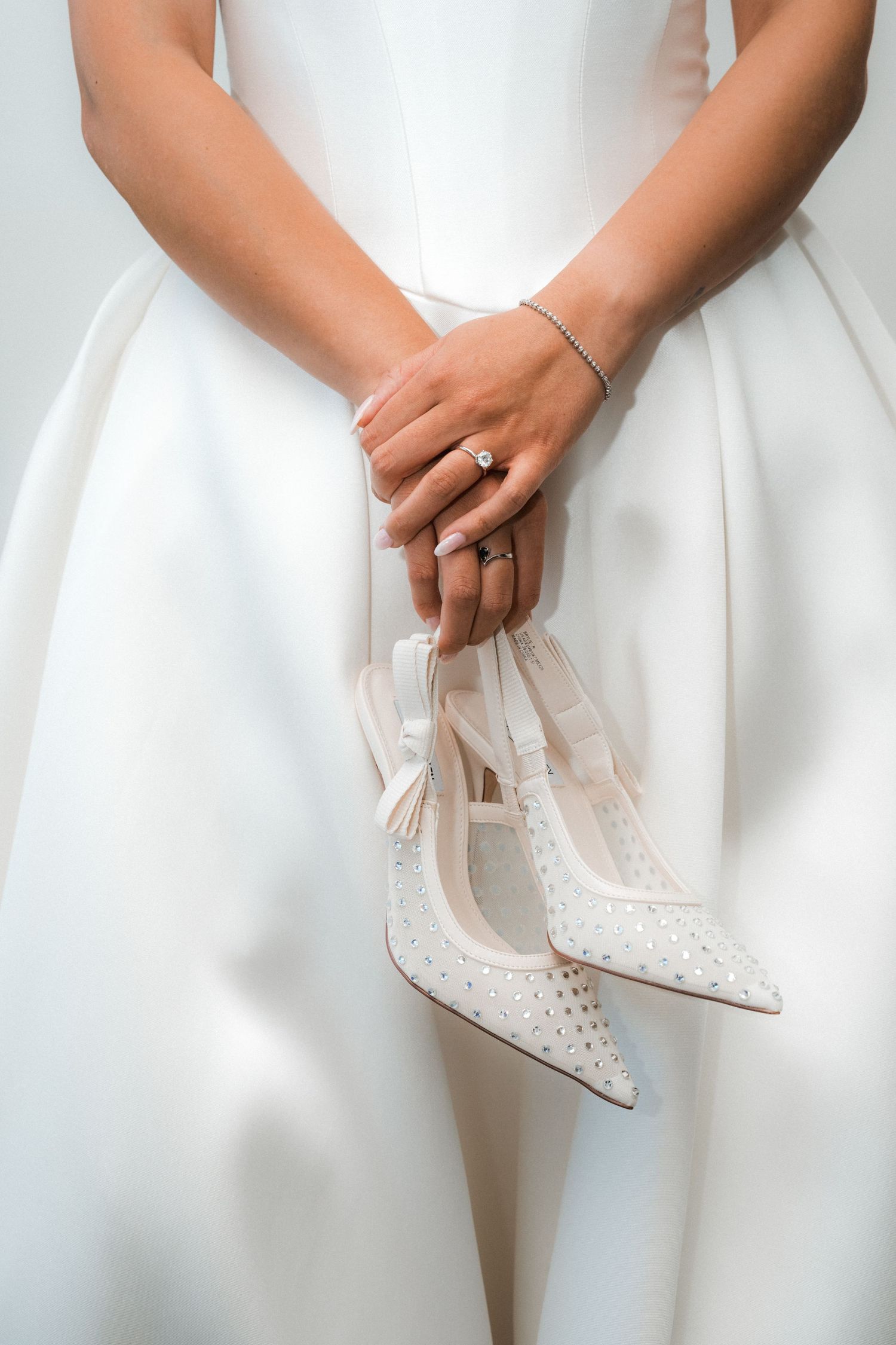 Bride's hands hold delicate white lace shoes against her wedding dress.