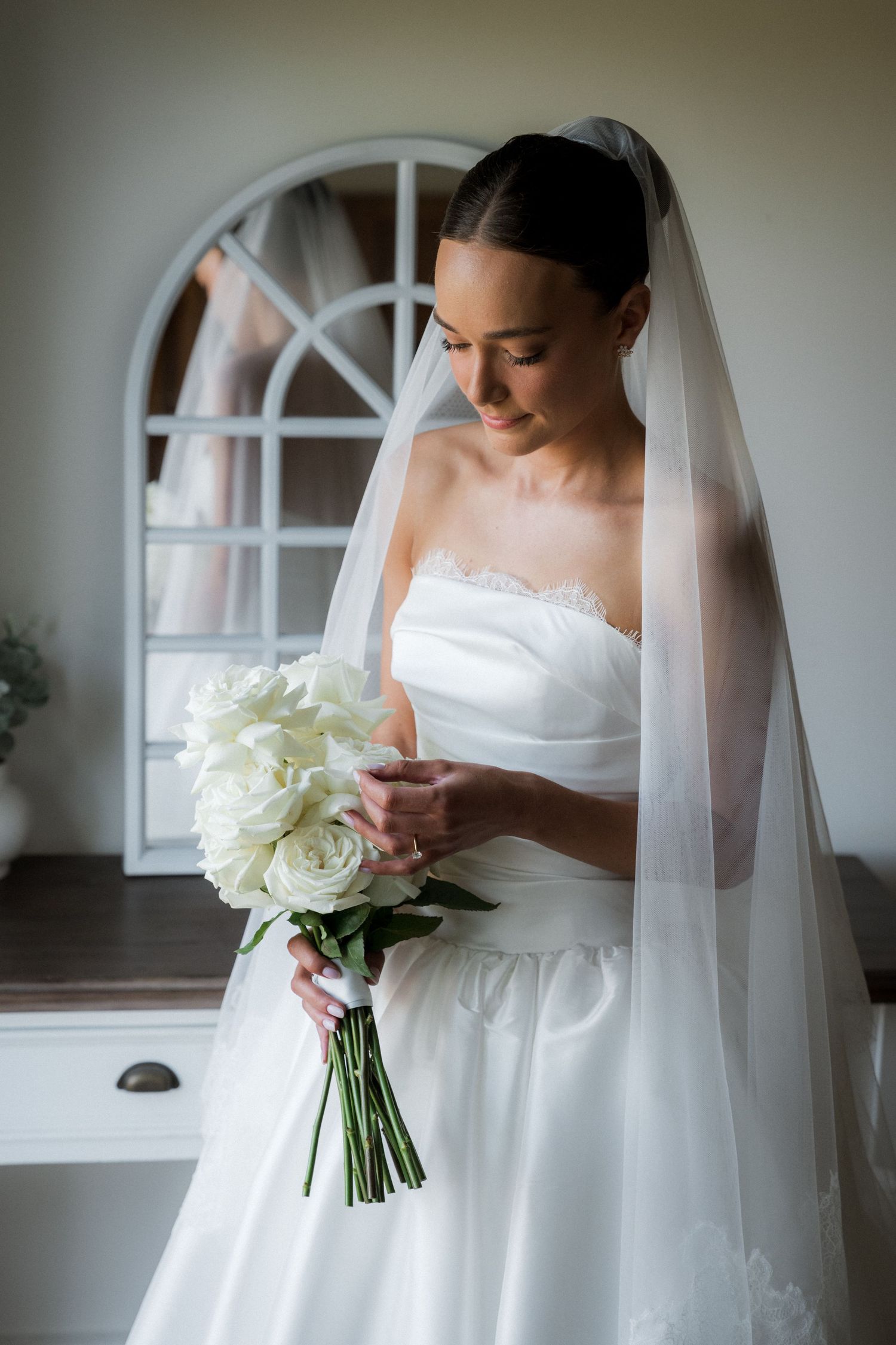 A bride in a white gown holds a bouquet of white flowers near an arched window.