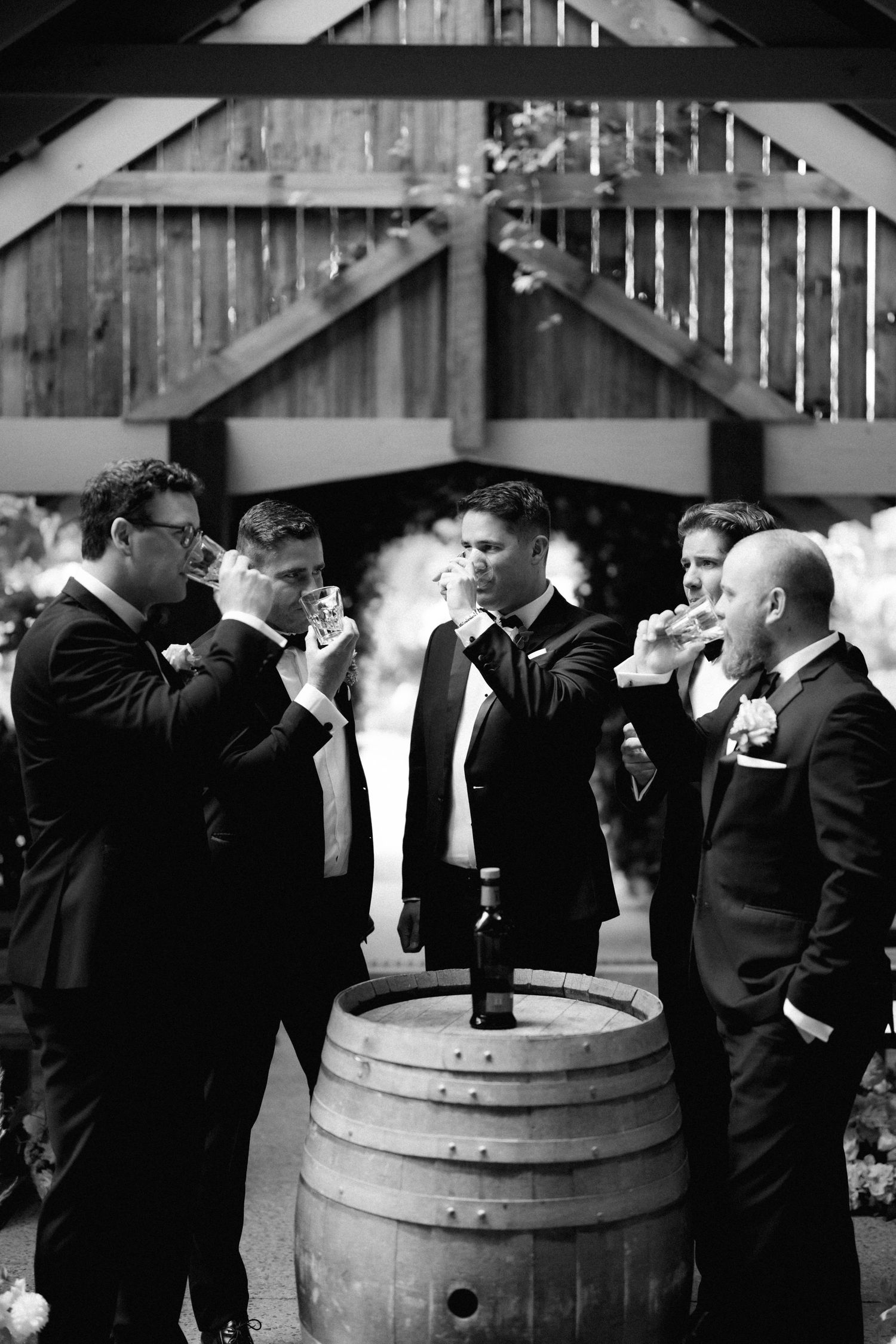 A black and white photo of groomsmen gathering around a wooden barrel outdoors, drinking and enjoying themselves.