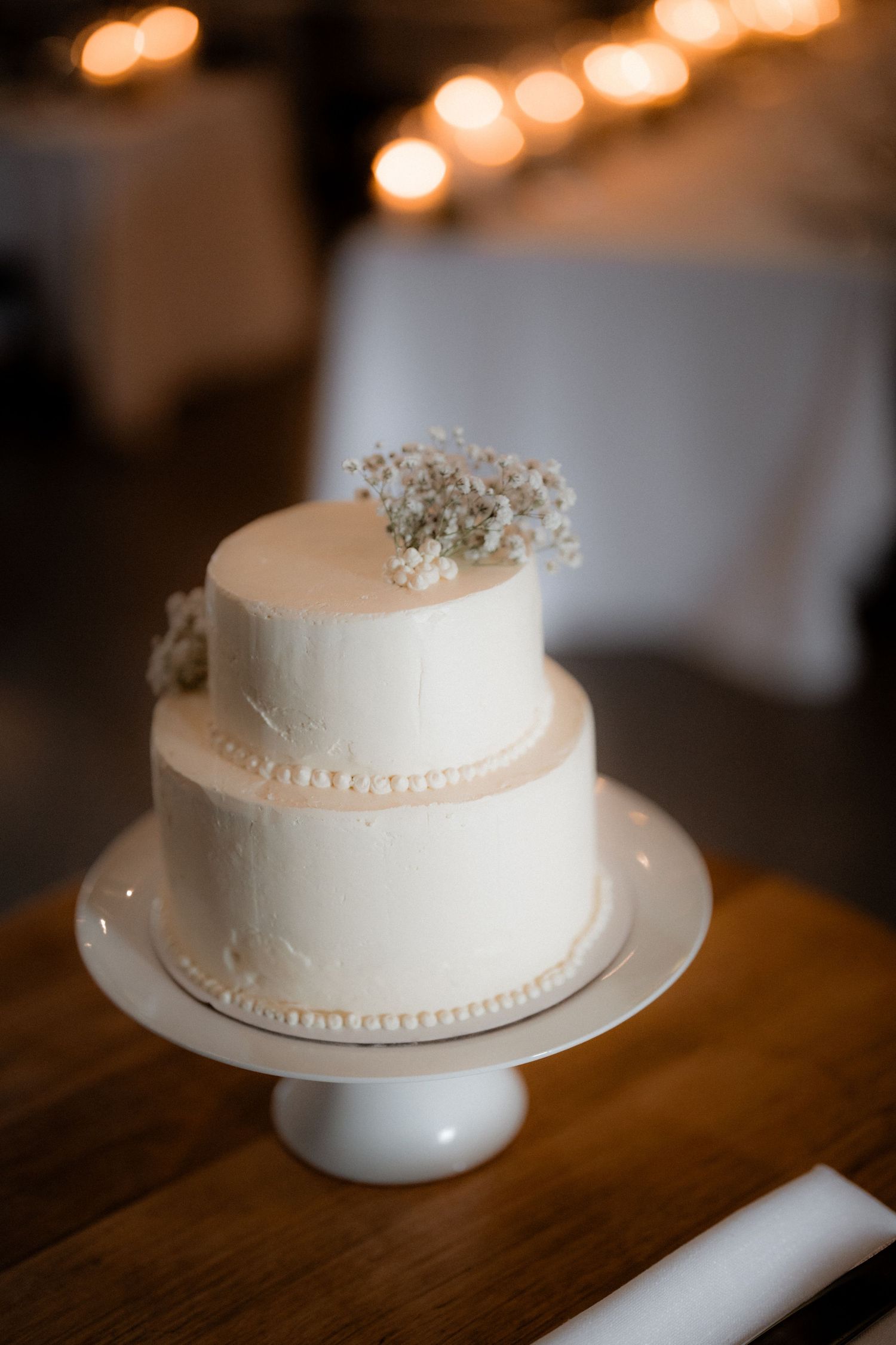 Elegant two-tiered white wedding cake with pearl details and baby's breath floral topper on a white pedestal stand.