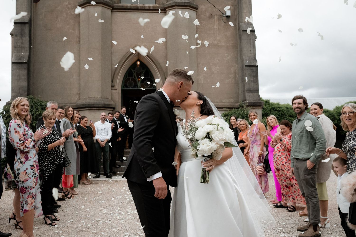 Two people in wedding attire share a kiss while guests throw confetti outside a church during their celebration.