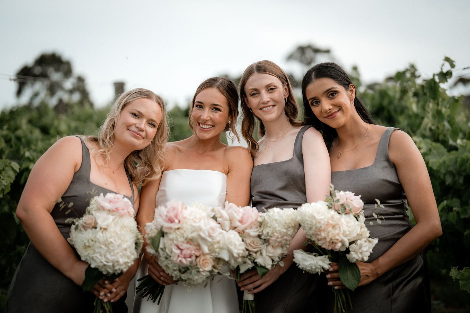 Bridal party in gray dresses holds elegant bouquets of white peonies at an outdoor vineyard wedding.