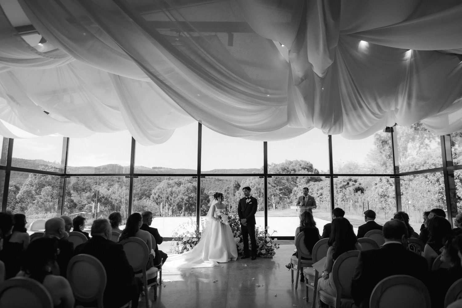 Bride and groom exchange vows during an indoor wedding ceremony with guests seated beneath draped white fabric and floor-to-ceiling windows.