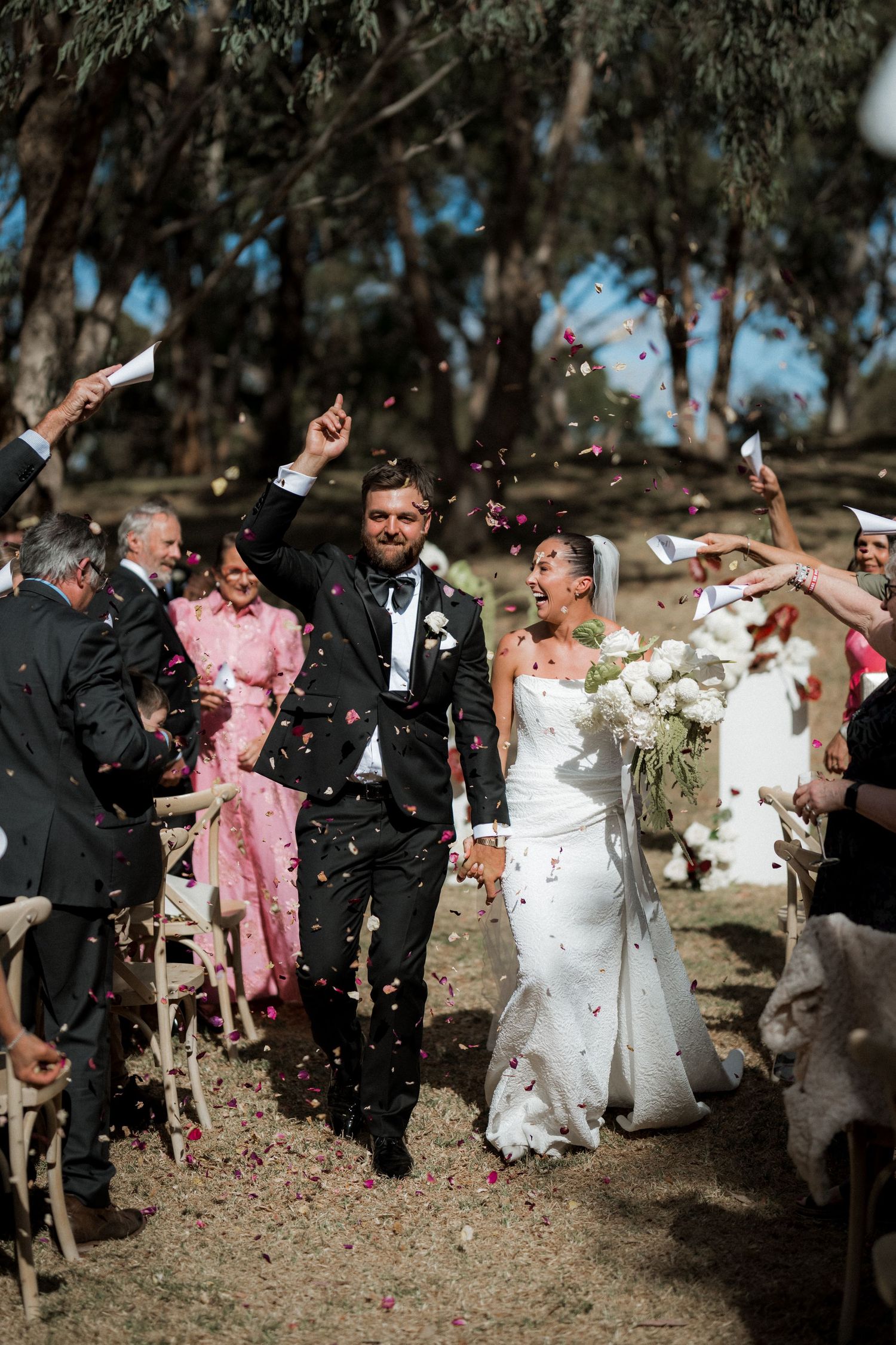 Newlyweds walk down the aisle as guests shower them with colorful confetti outdoors.