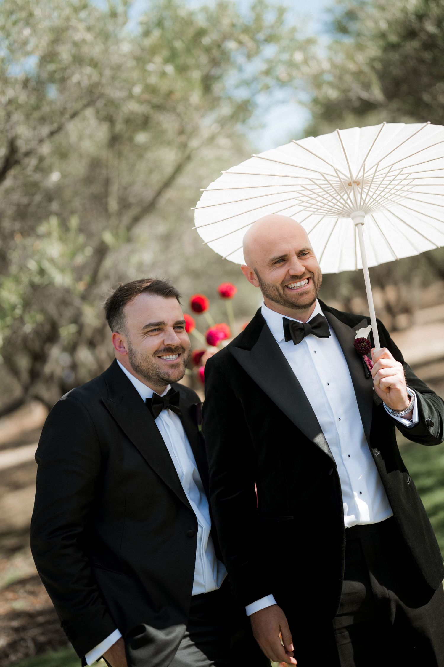 Groomsmen in tuxedos playfully pose with white parasol outdoors.