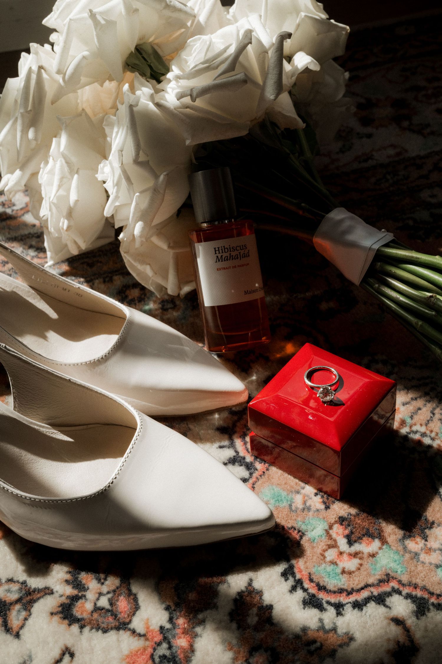 White bridal shoes and bouquet arranged with red gift boxes on a patterned carpet.