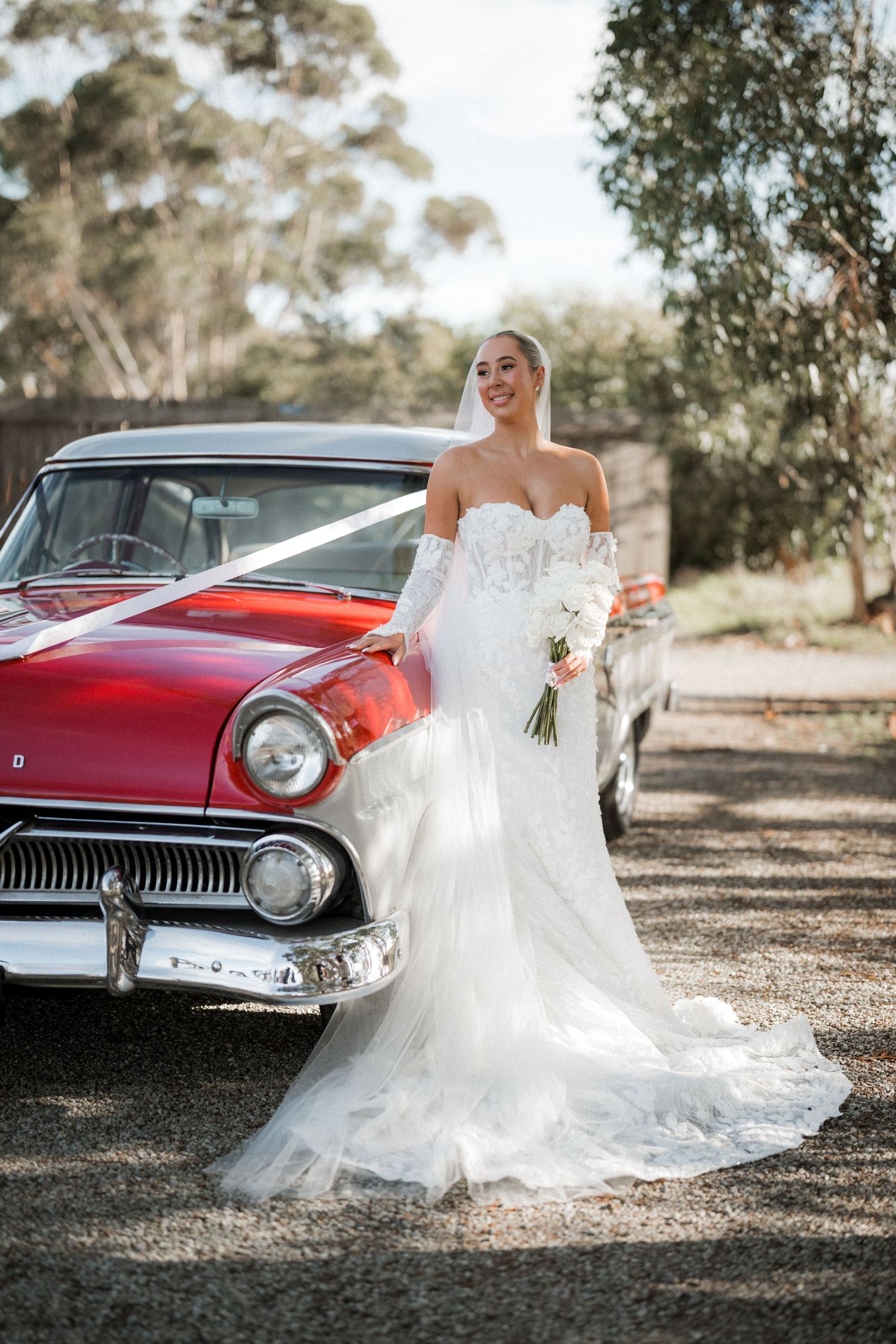 A bride in a flowing white dress poses next to a vintage red car in a rustic outdoor setting.