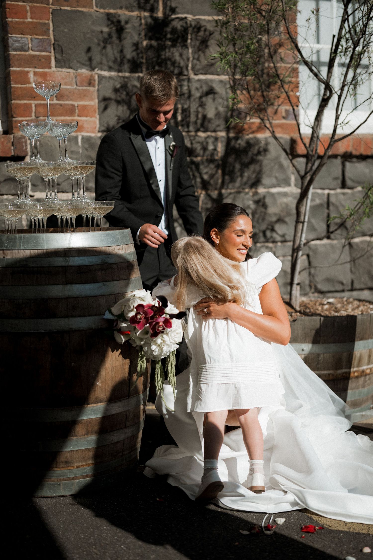 A series of candid wedding photos showing a bride and flower girl in sunlit moments by a brick wall.