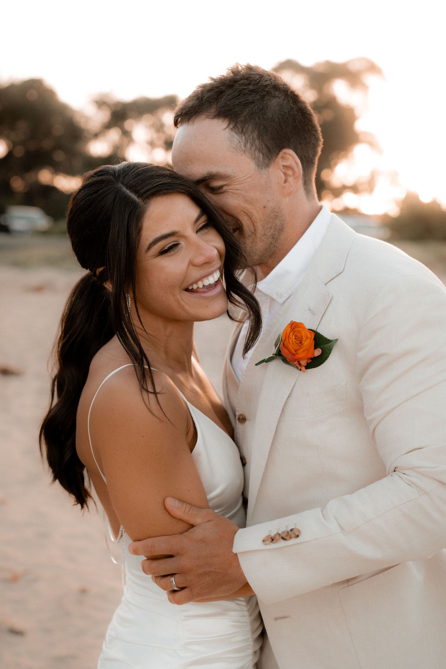 A joyful couple shares an intimate moment at sunset, with the groom wearing a white suit and orange boutonniere.
