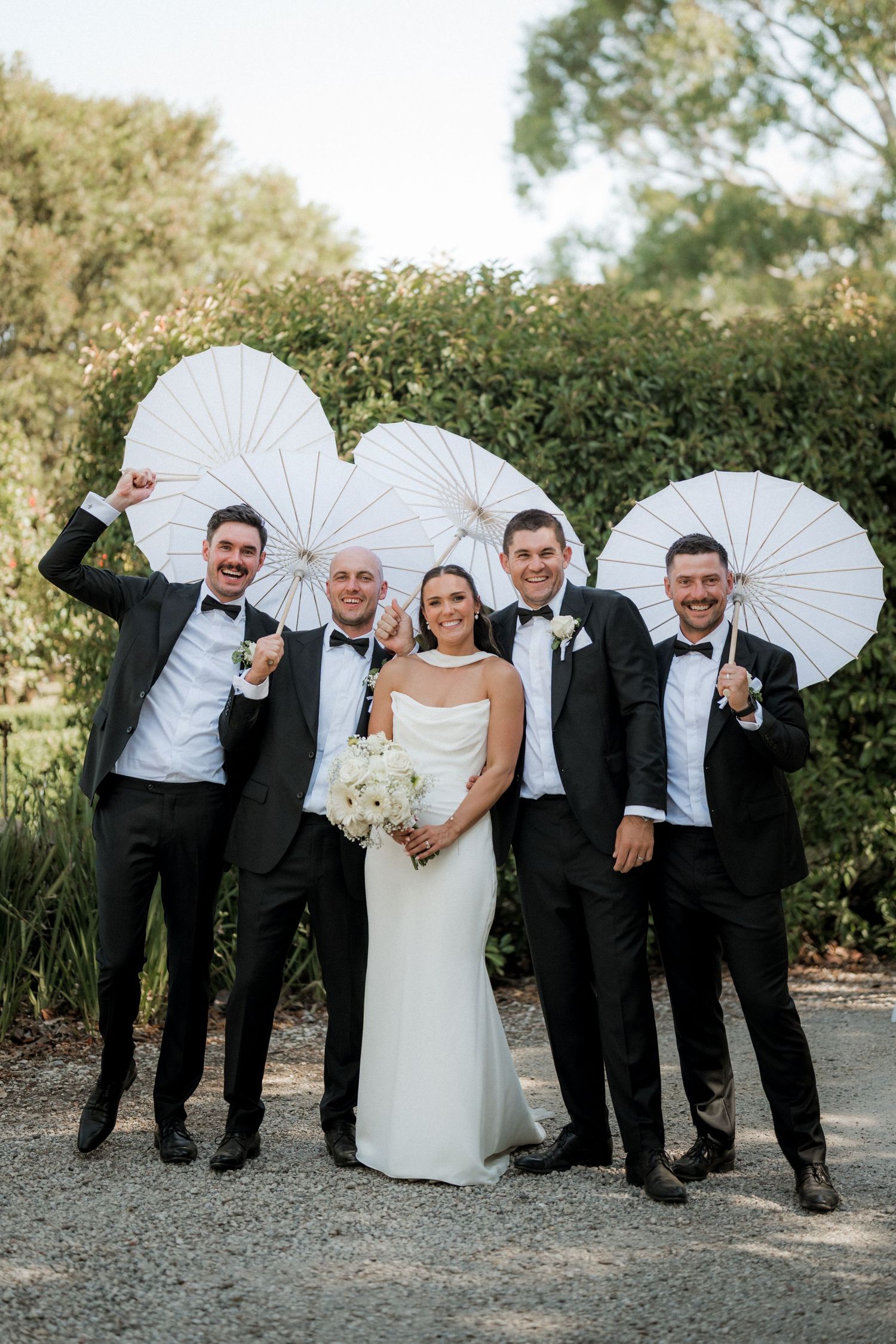 Wedding party poses playfully with white parasols against garden backdrop.