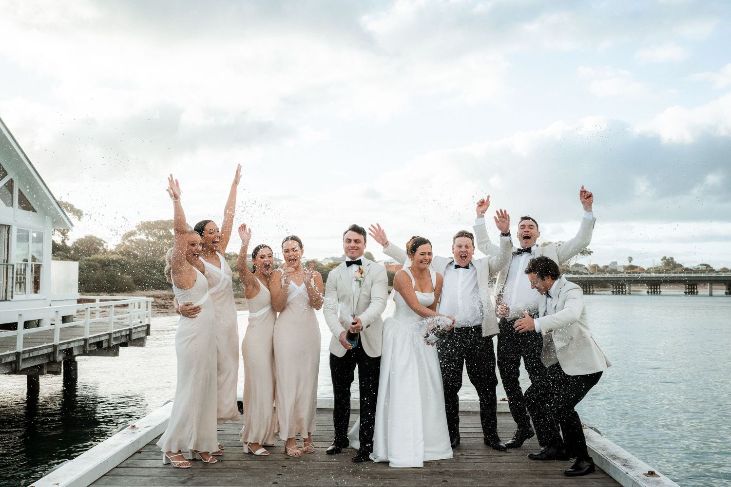 Wedding party celebrates joyfully on a waterfront dock with boats and buildings in background at sunset.