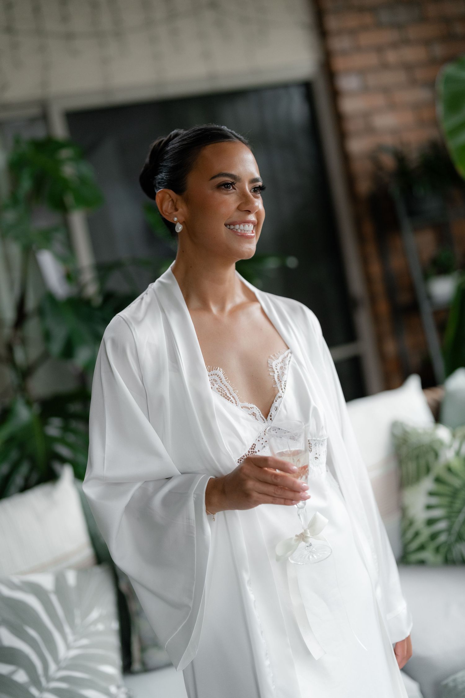 A bride in a white silk robe holds a glass while getting ready in an elegant room with brick walls and plants.