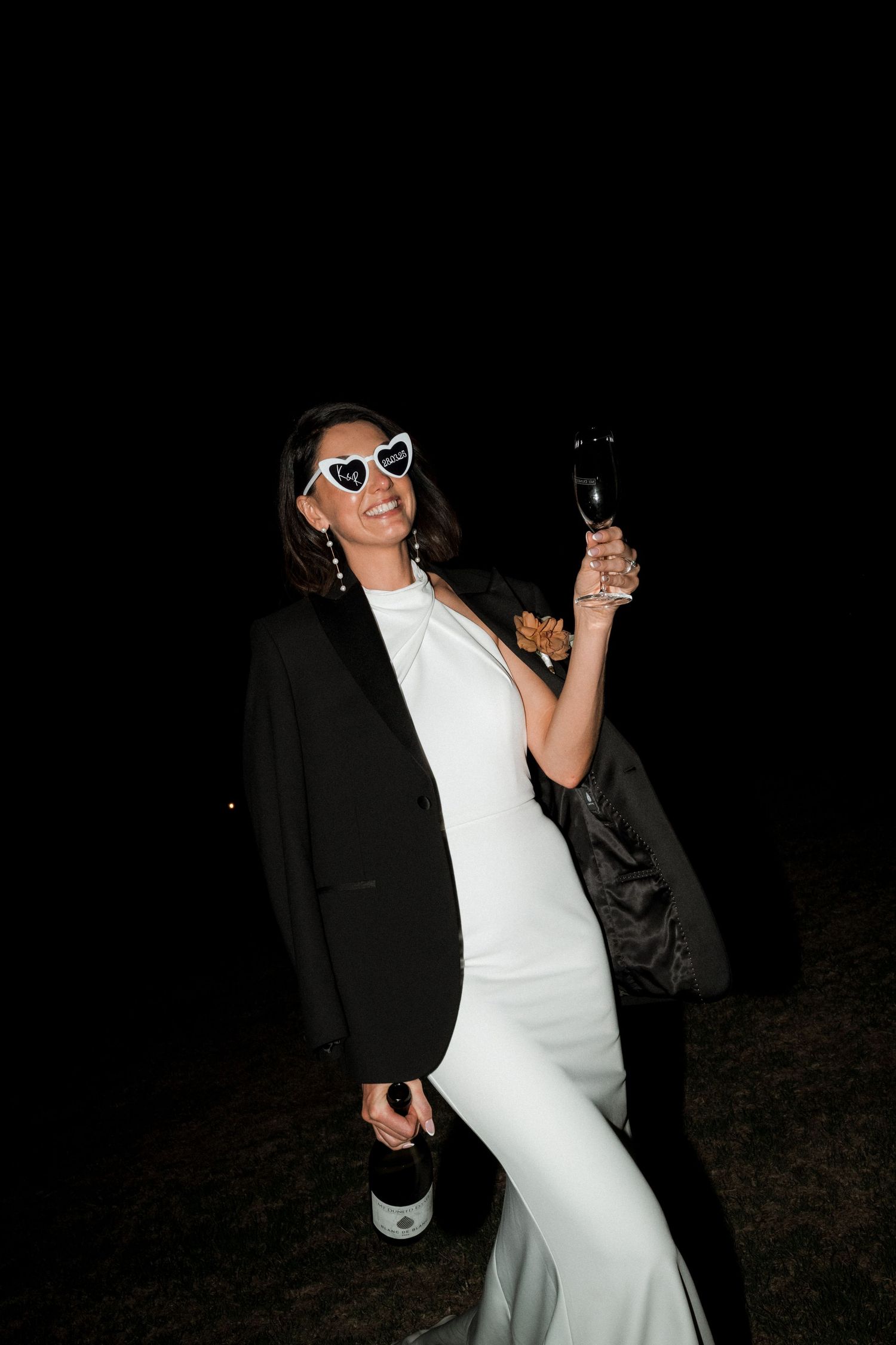 A person in a black blazer and white dress holds wine glasses at night while wearing heart-shaped sunglasses.