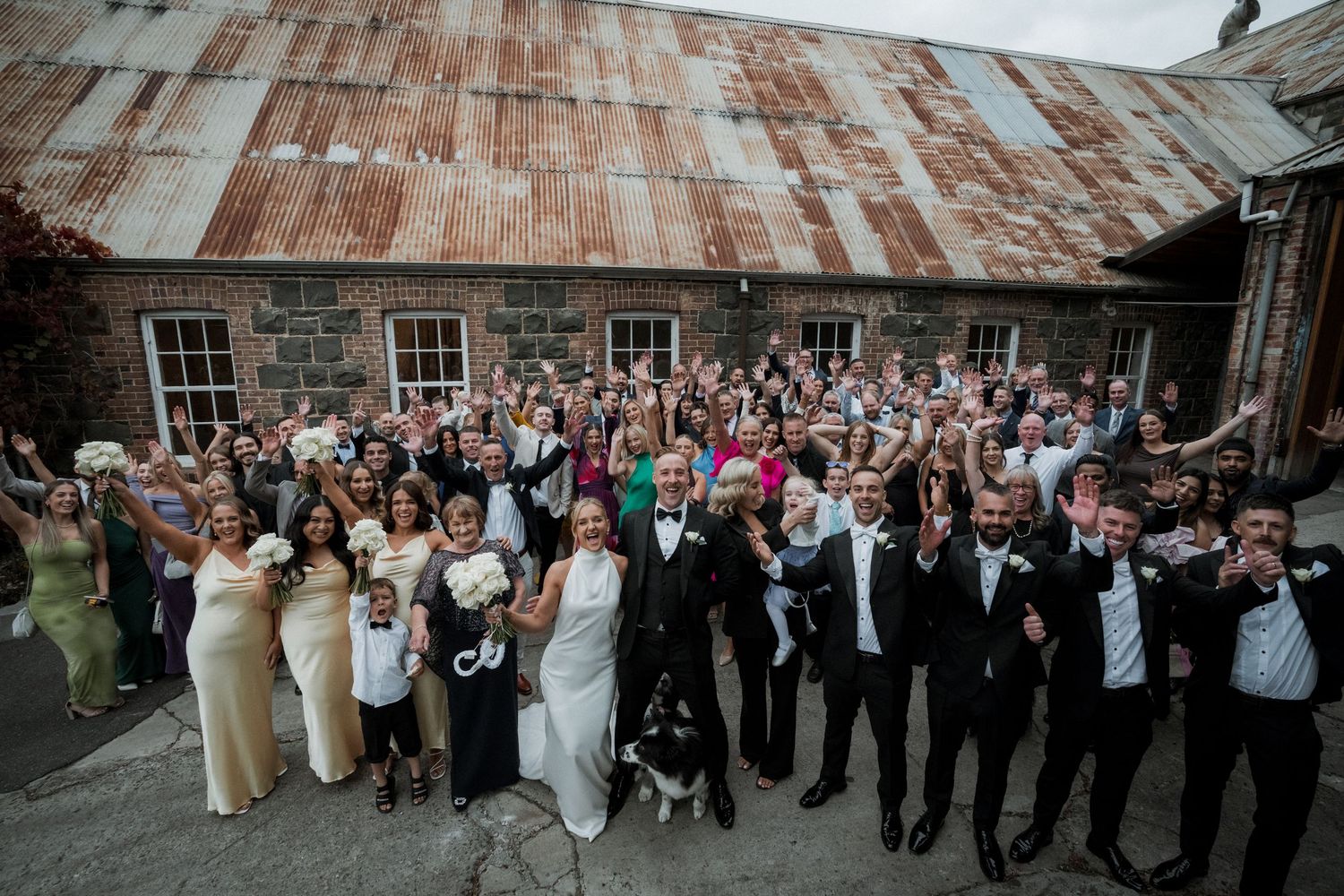 Large wedding party and guests pose together for group photo outside rustic barn with weathered metal roof.