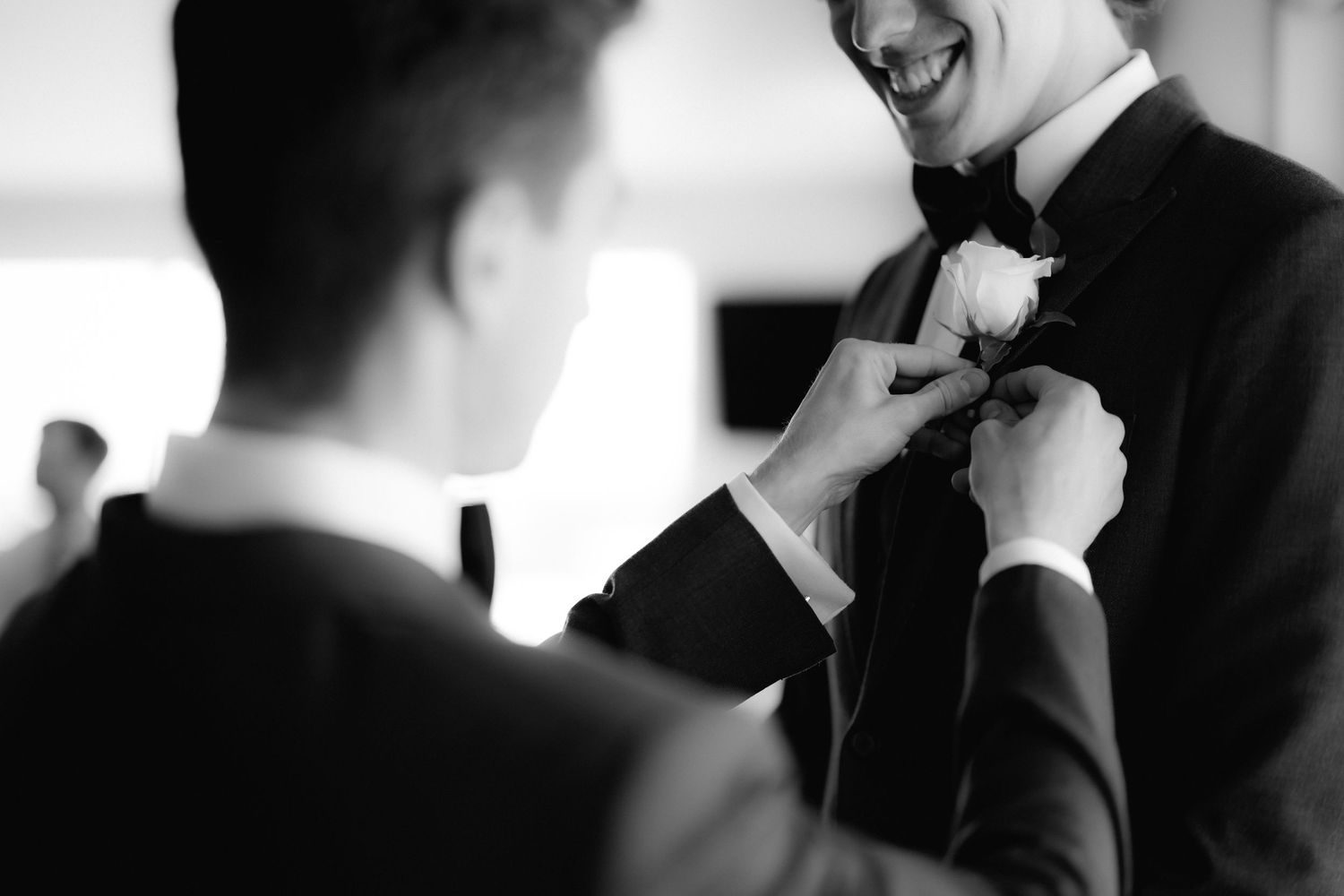 A groomsman helps adjust a wedding boutonniere on the lapel of a formal black tuxedo in black and white.
