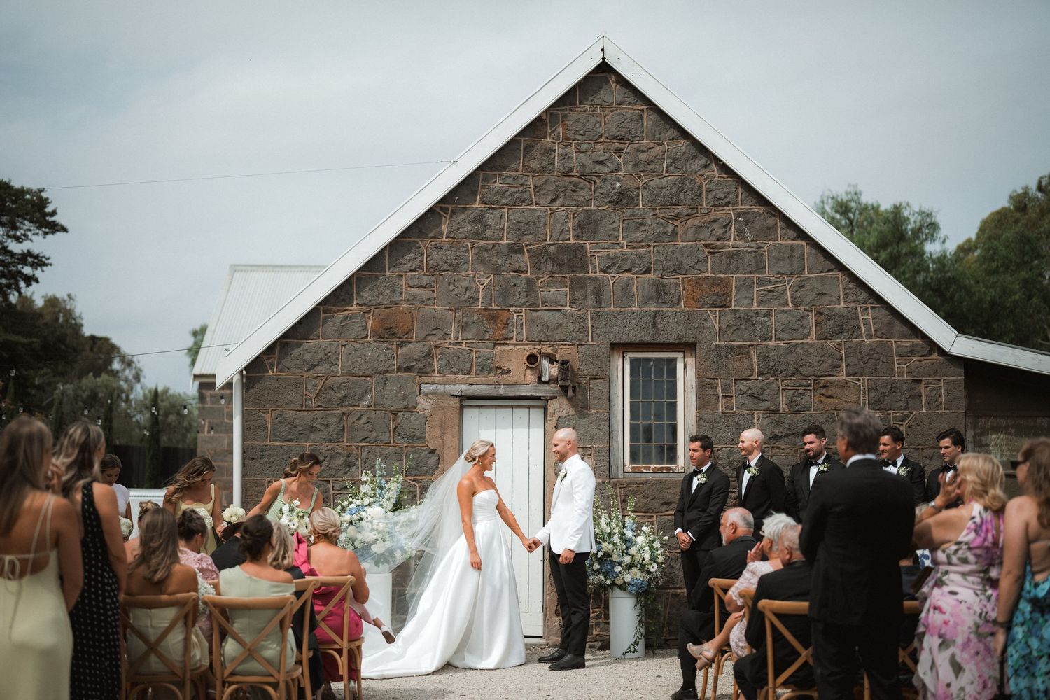 A scenic outdoor wedding ceremony takes place outside a rustic stone church building with guests seated on both sides.