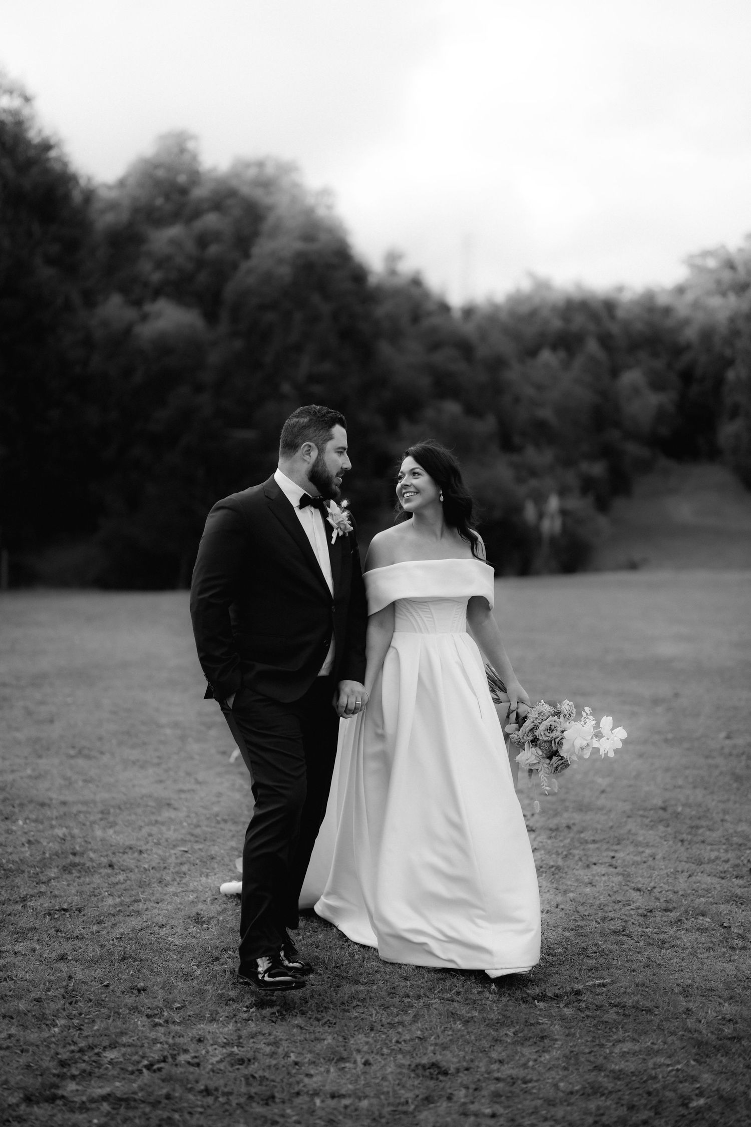 A bride and groom share a romantic moment on a grassy field with trees in the background, captured in black and white.