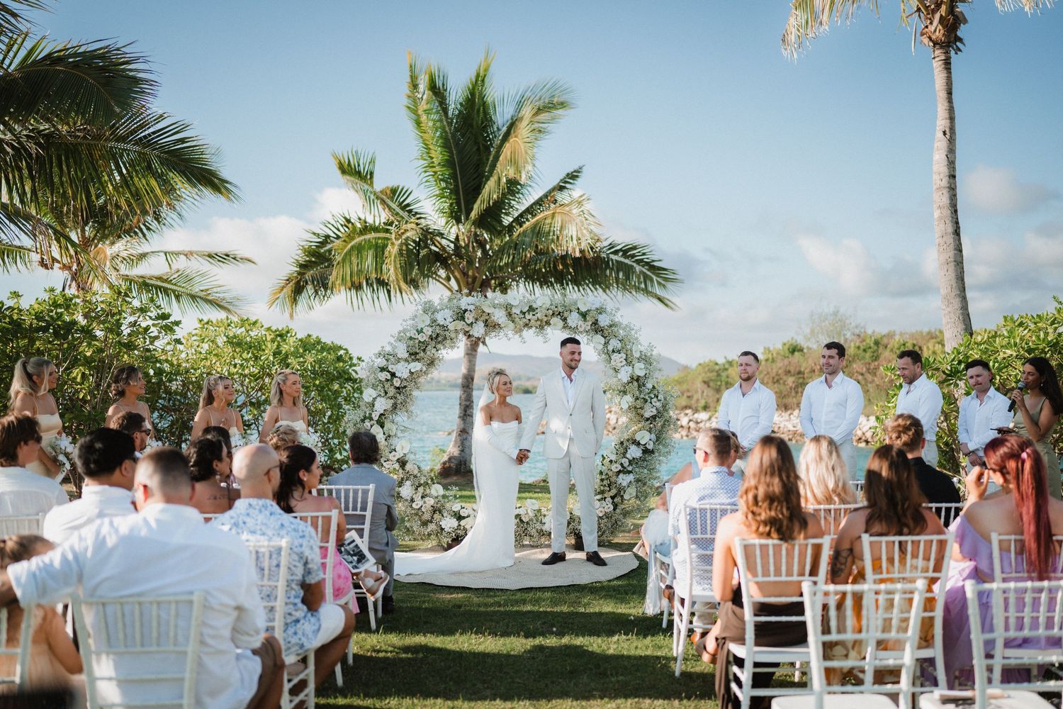 Outdoor wedding ceremony setup with white chairs, floral arch and palm trees overlooking the ocean.