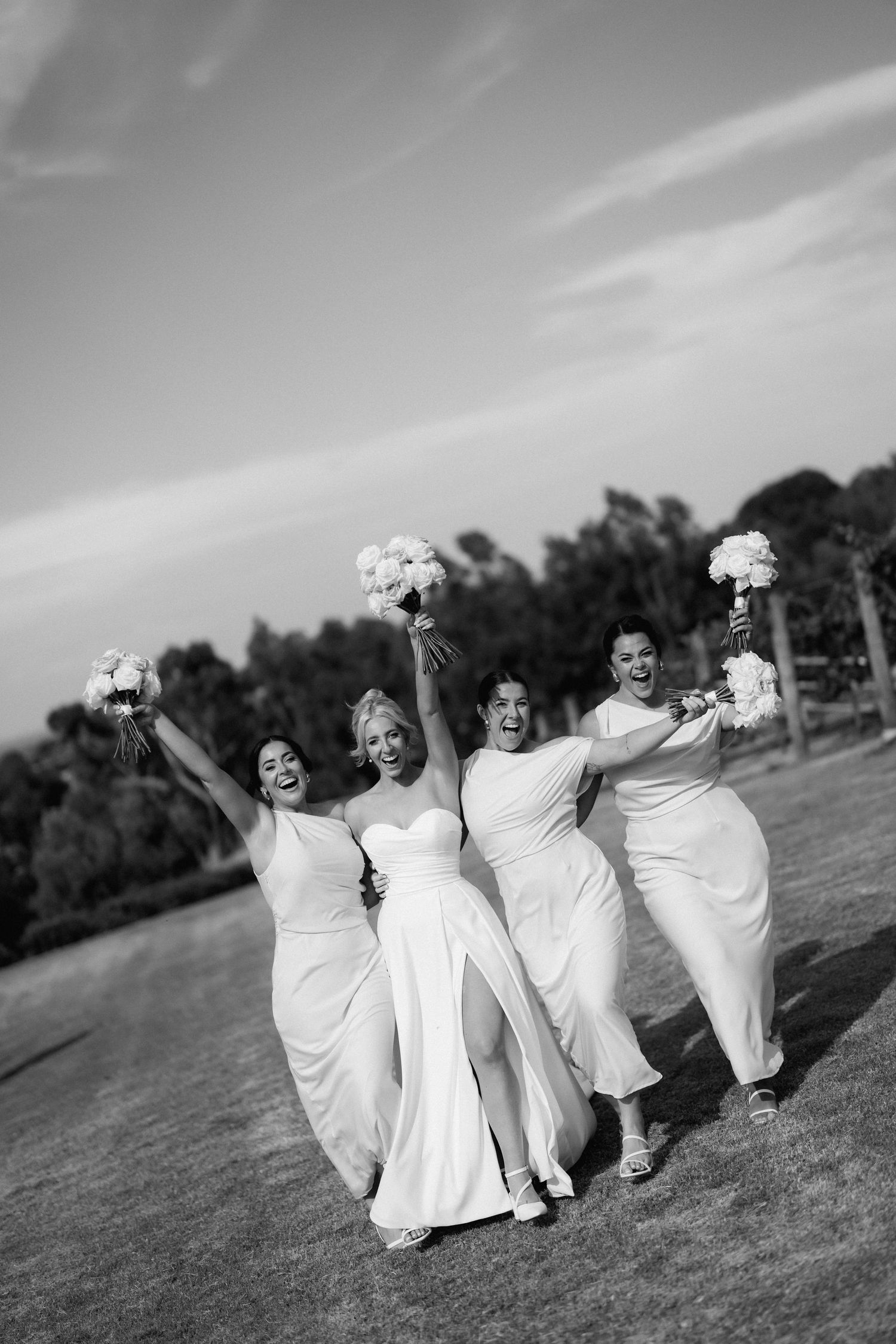 Bridesmaids in white dresses celebrate joyfully while holding bouquets on a grassy hill.