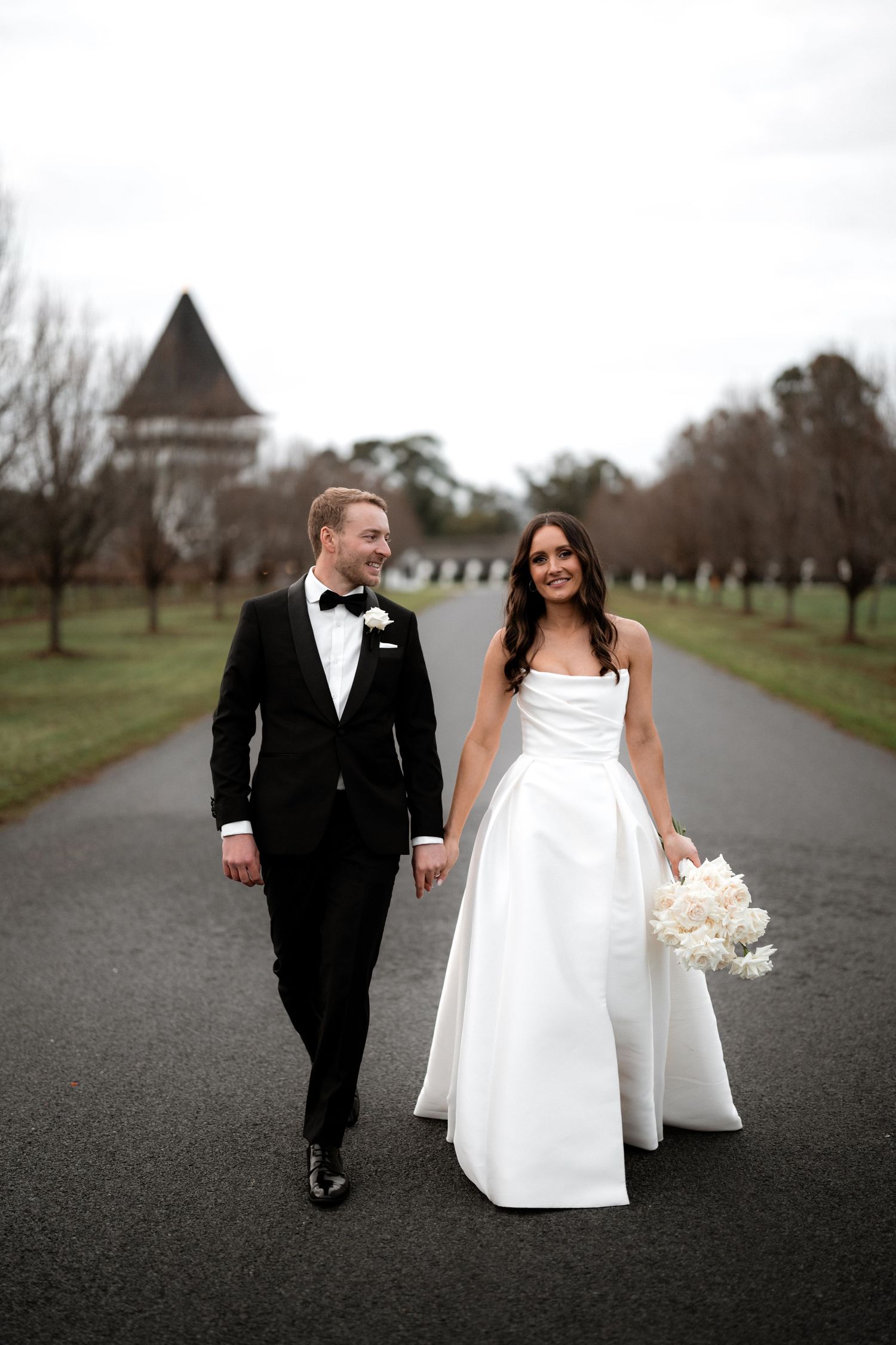 Couple walks down tree-lined path in wedding attire with historic building in background.