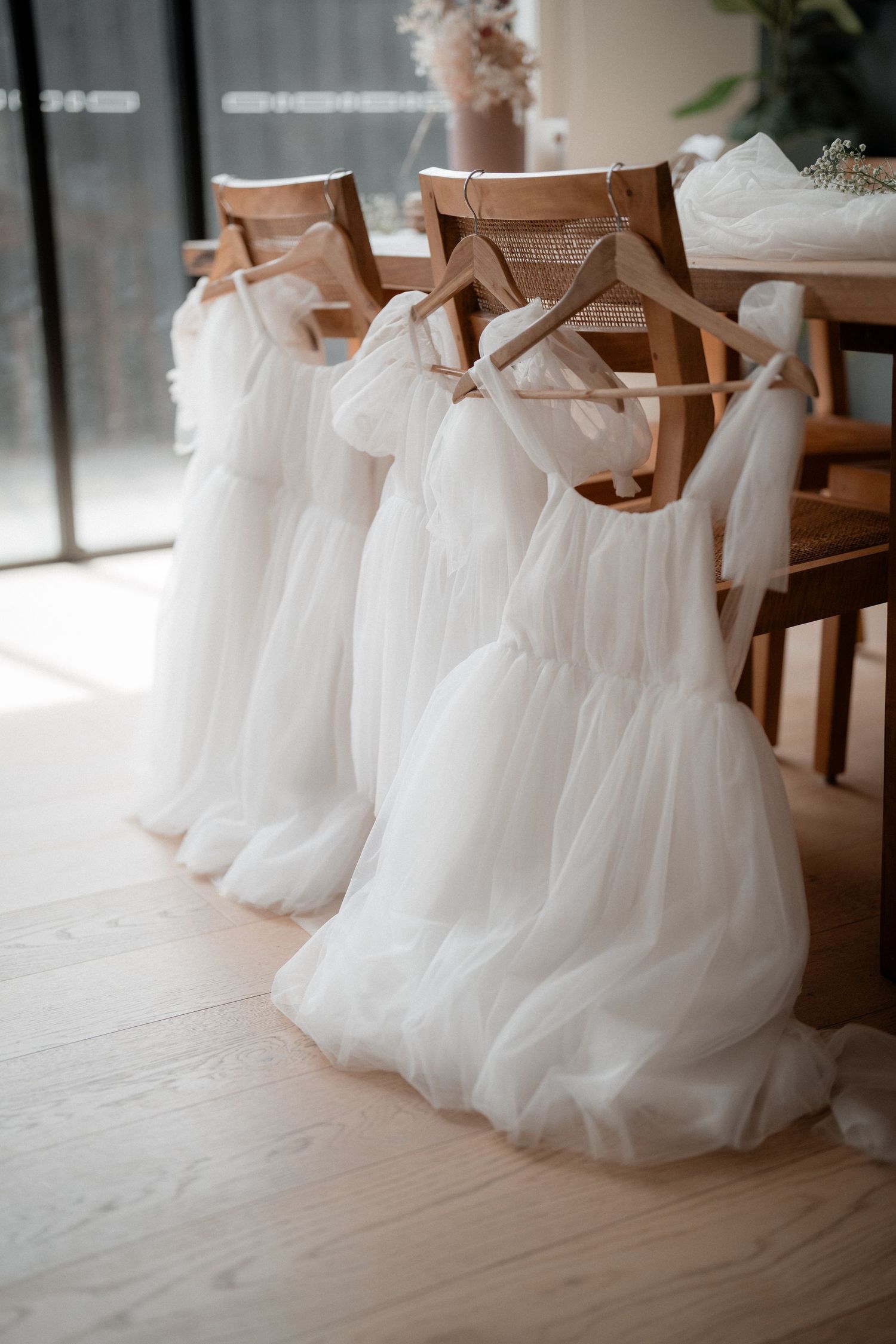 White tulle flower girl dresses hanging on wooden hangers in natural light against wooden floor.