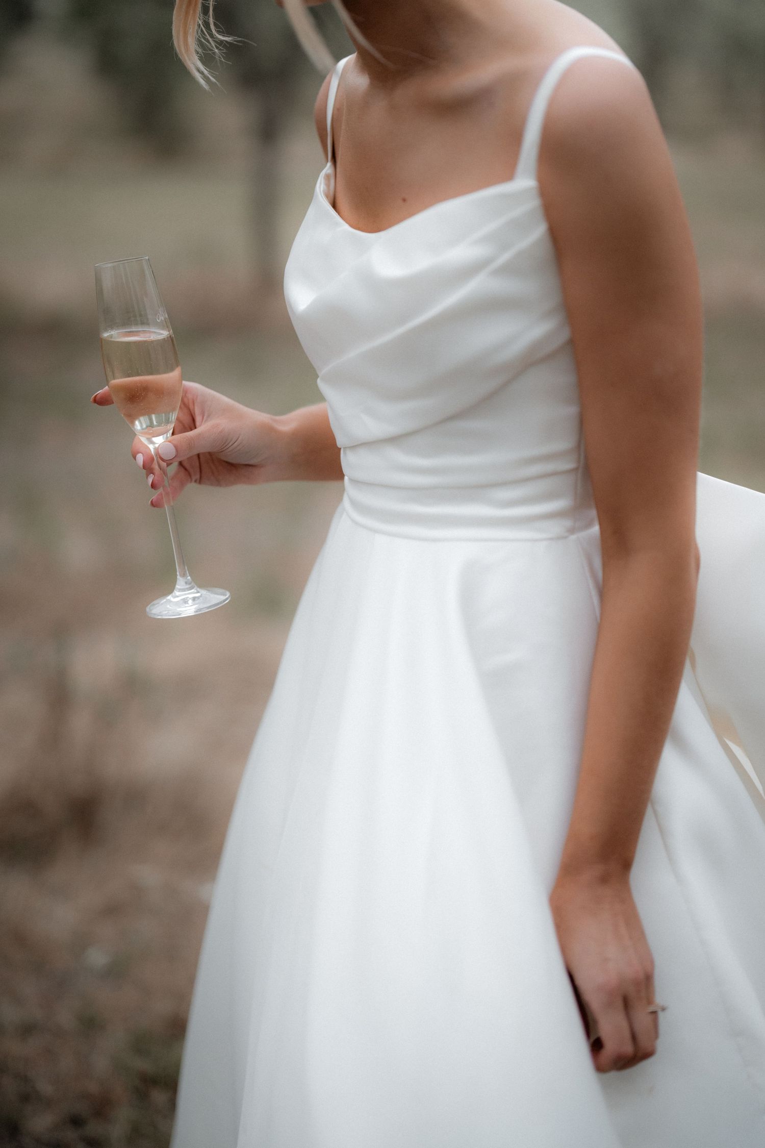 Elegant white satin wedding dress with spaghetti straps shown with champagne glass in hand.