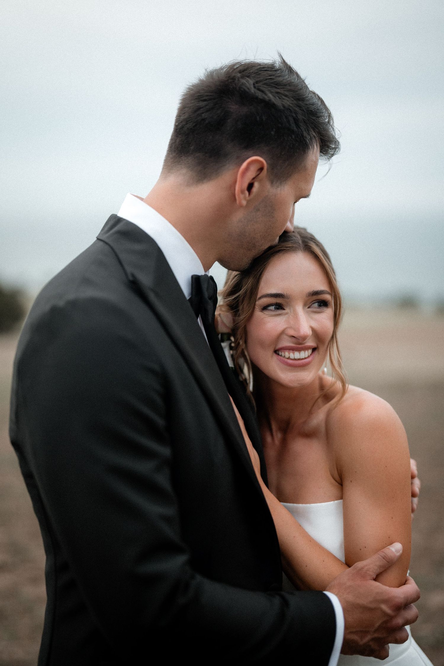A tender moment between newlyweds as they embrace on their wedding day against an overcast sky.