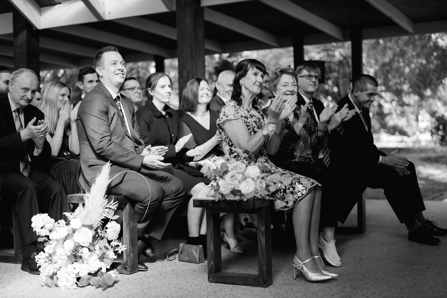 A black and white photograph of wedding guests sitting in a row and clapping during an outdoor celebration.