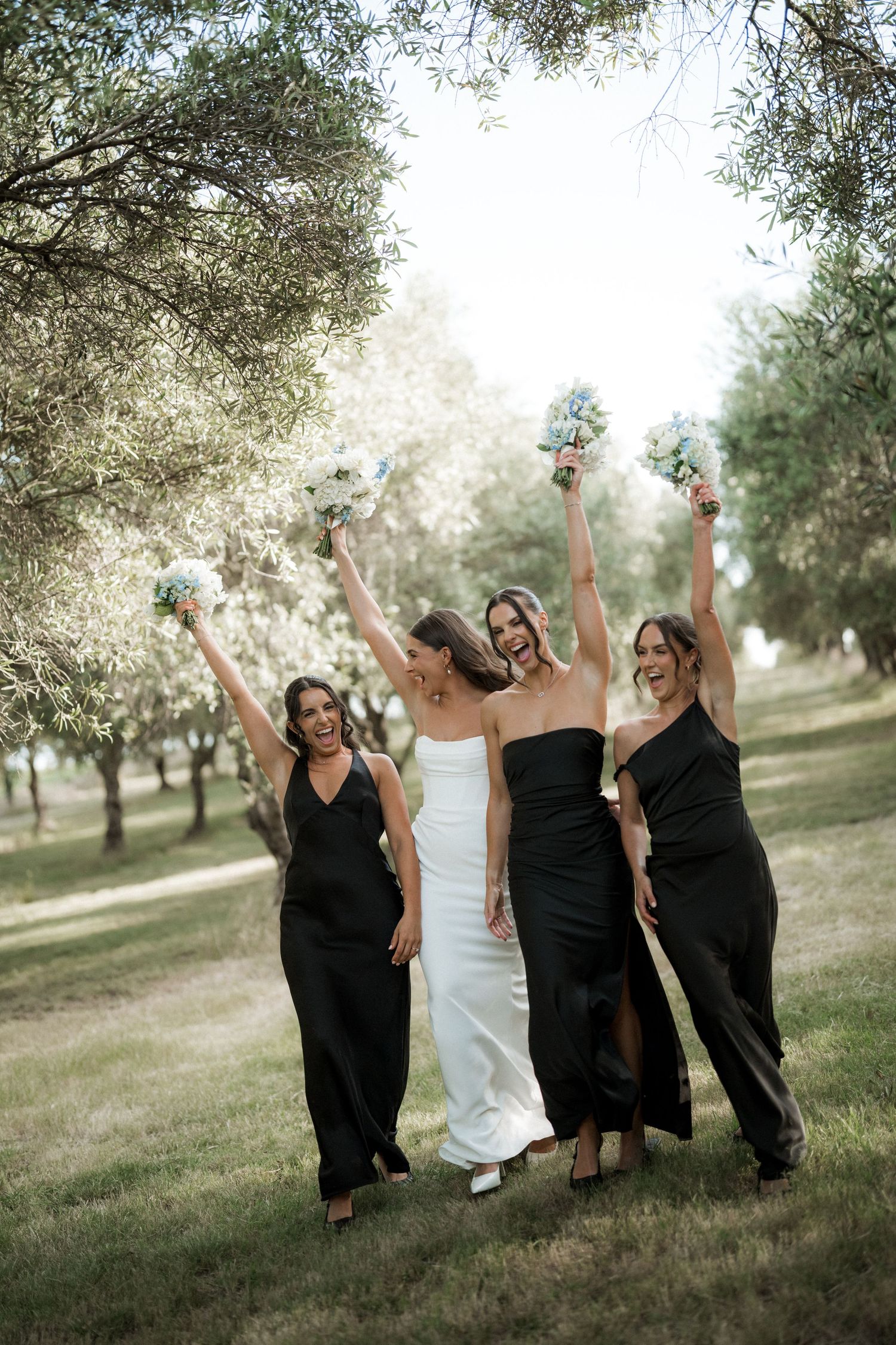 Bridal party in black dresses celebrates joyfully among olive trees in outdoor setting.