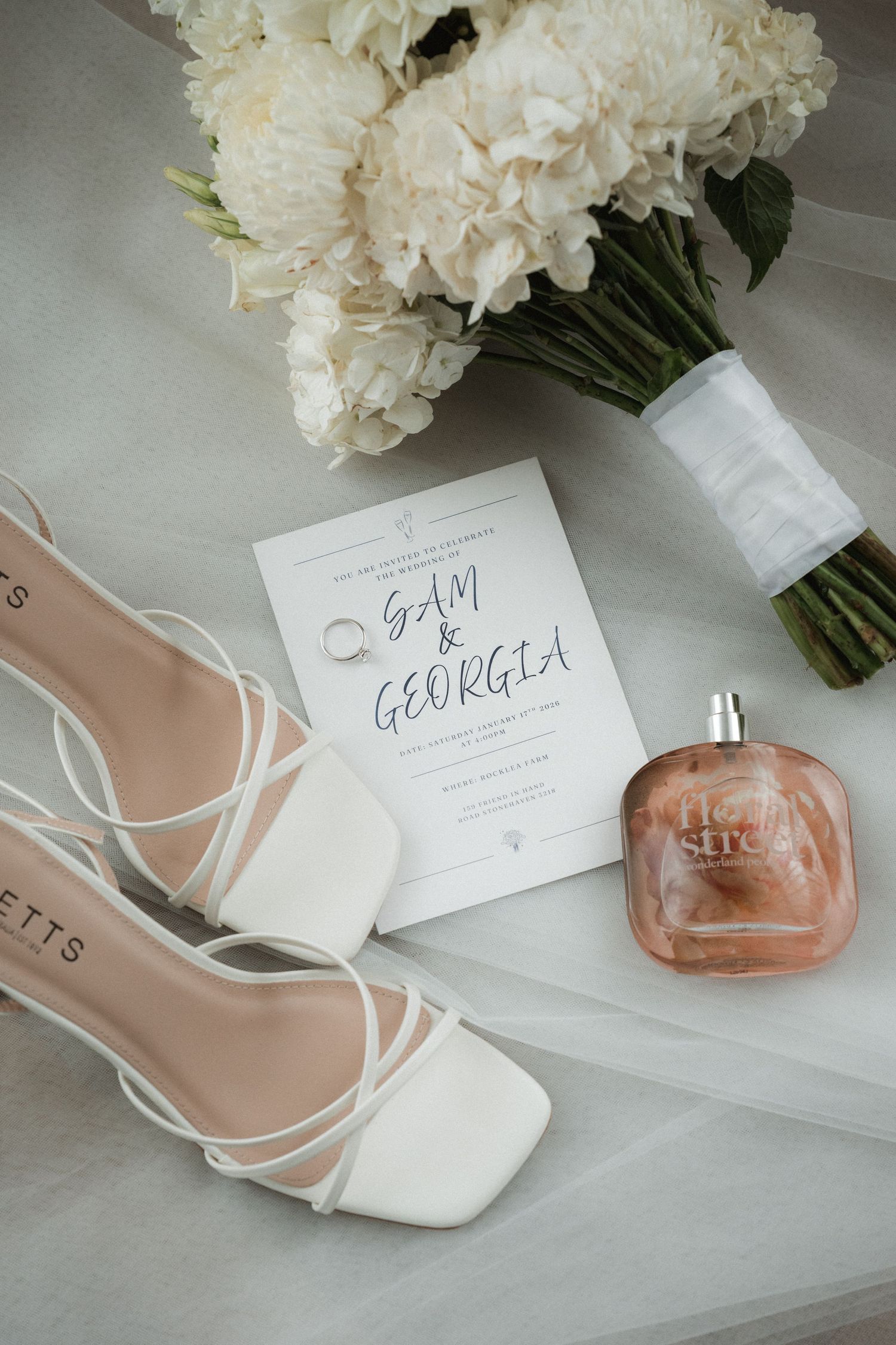 Wedding details featuring white bridal shoes, perfume bottles, and elegant white floral bouquets arranged on a light surface.