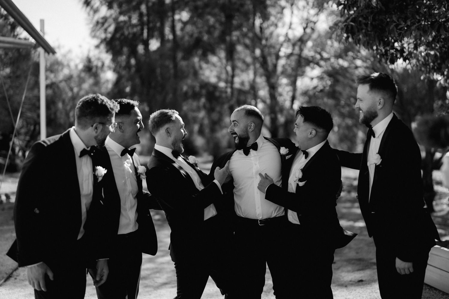 Groomsmen in black tuxedos pose together outdoors in a candid black and white photo.