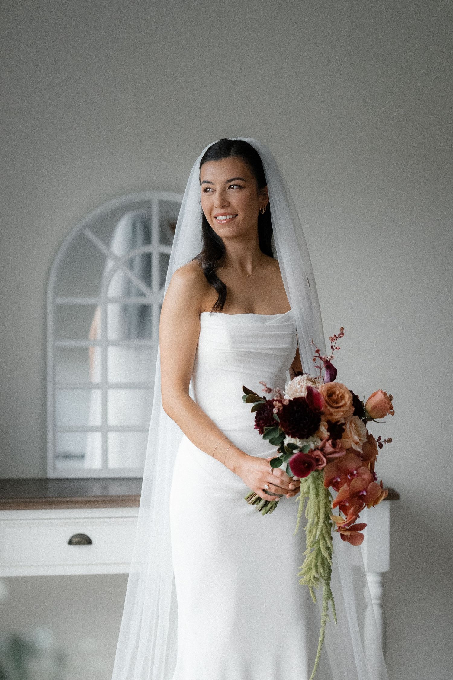 Bride in white strapless gown and veil holding a cascading bouquet of burgundy and peach flowers indoors.