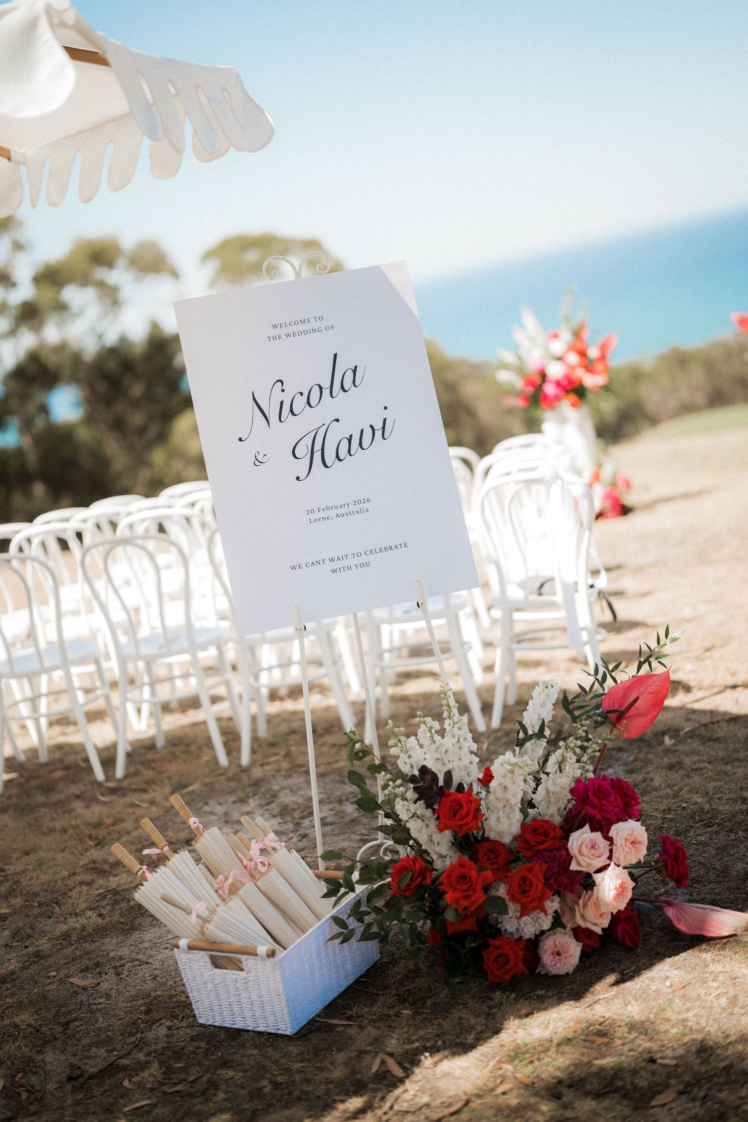 Outdoor wedding ceremony setup with white chairs, welcome sign for Nicola & Havi, rolled programs, and red floral arrangement by the sea.