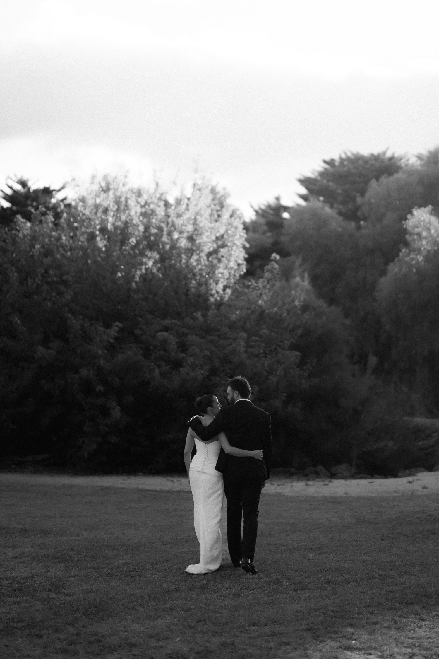 Black and white photo of bride and groom embracing while gazing at trees in a field.