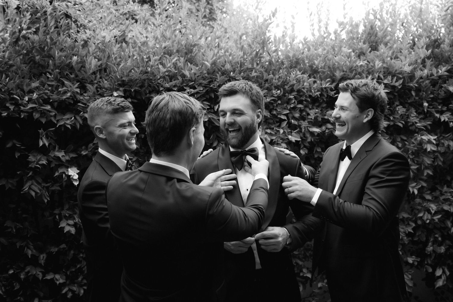 Groomsmen in tuxedos adjusting the grooms bow tie outdoors in a black and white wedding photo.