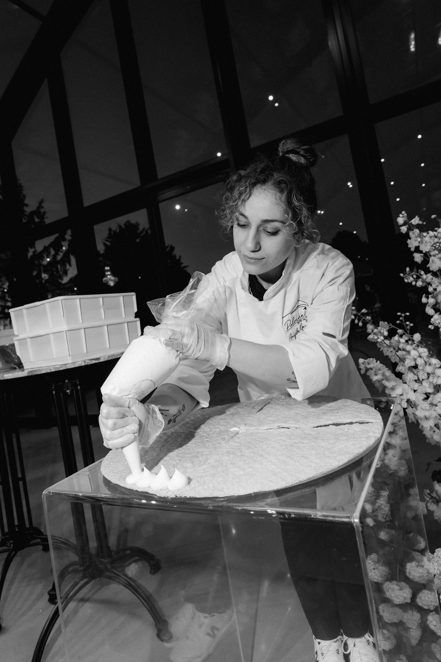 Pastry chef in a white coat pipes whipped cream from a piping bag onto a large round cake base at an elegant event.