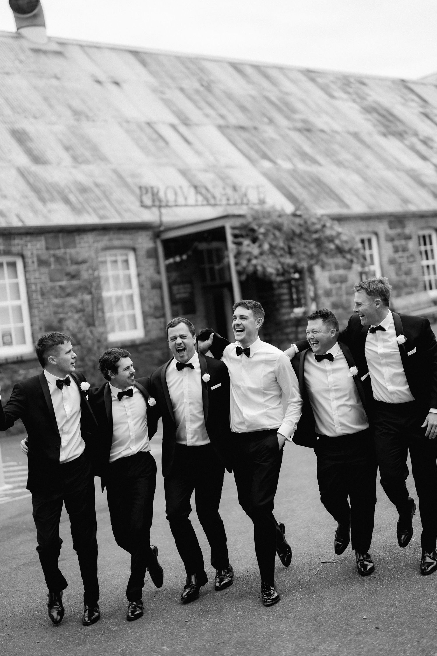 Groomsmen in black tuxedos and bow ties perform a synchronized jumping pose outside a brick building.