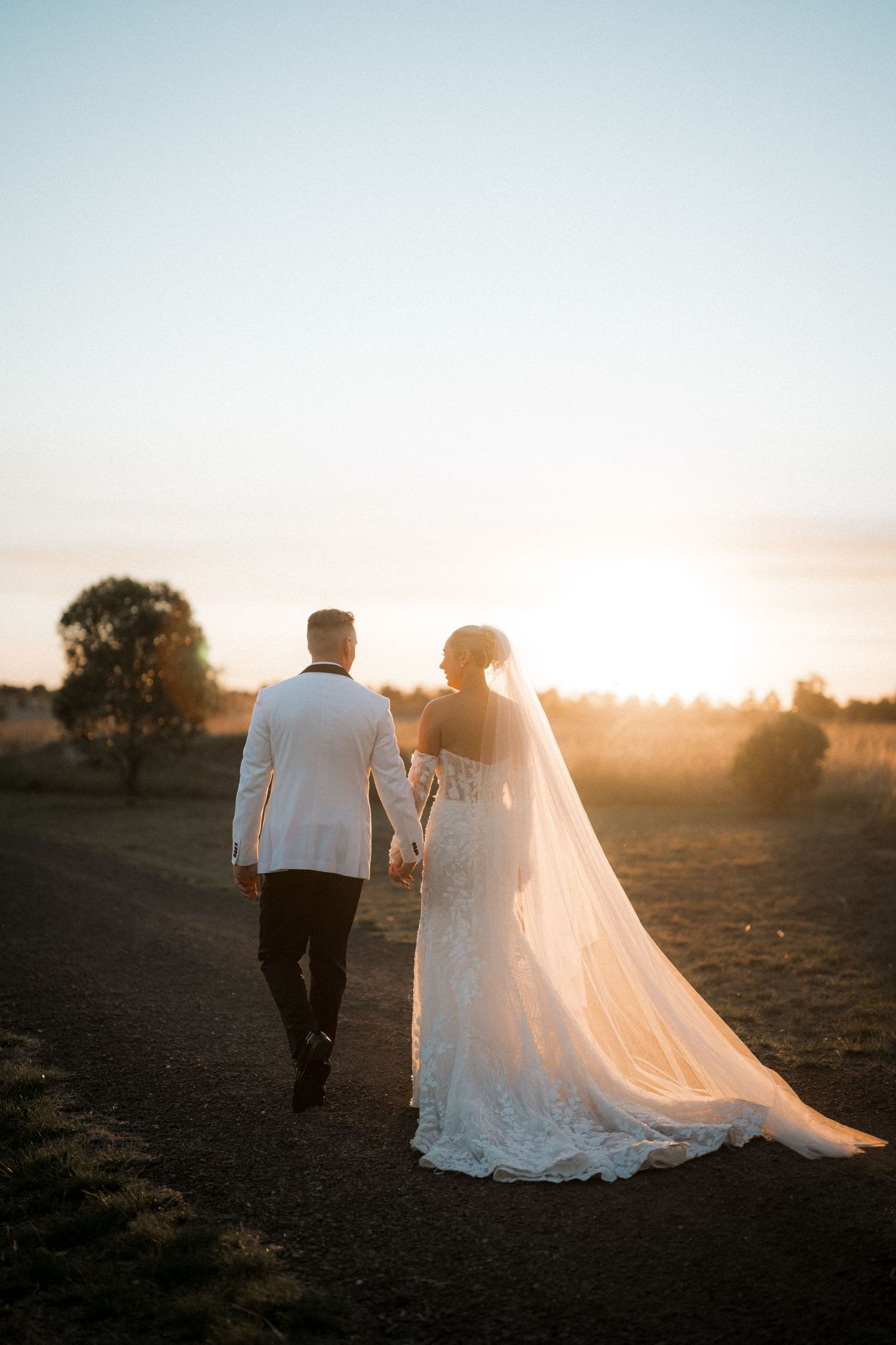 Newlywed couple walking hand in hand during golden sunset with bride wearing flowing lace gown and long veil.