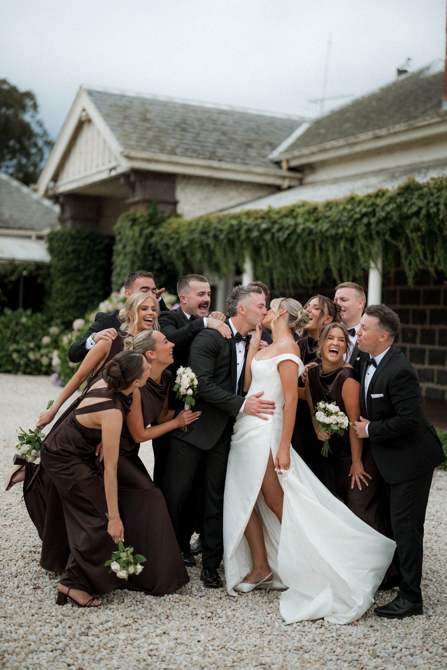 Wedding party poses for fun group photo on gravel path in formal attire.