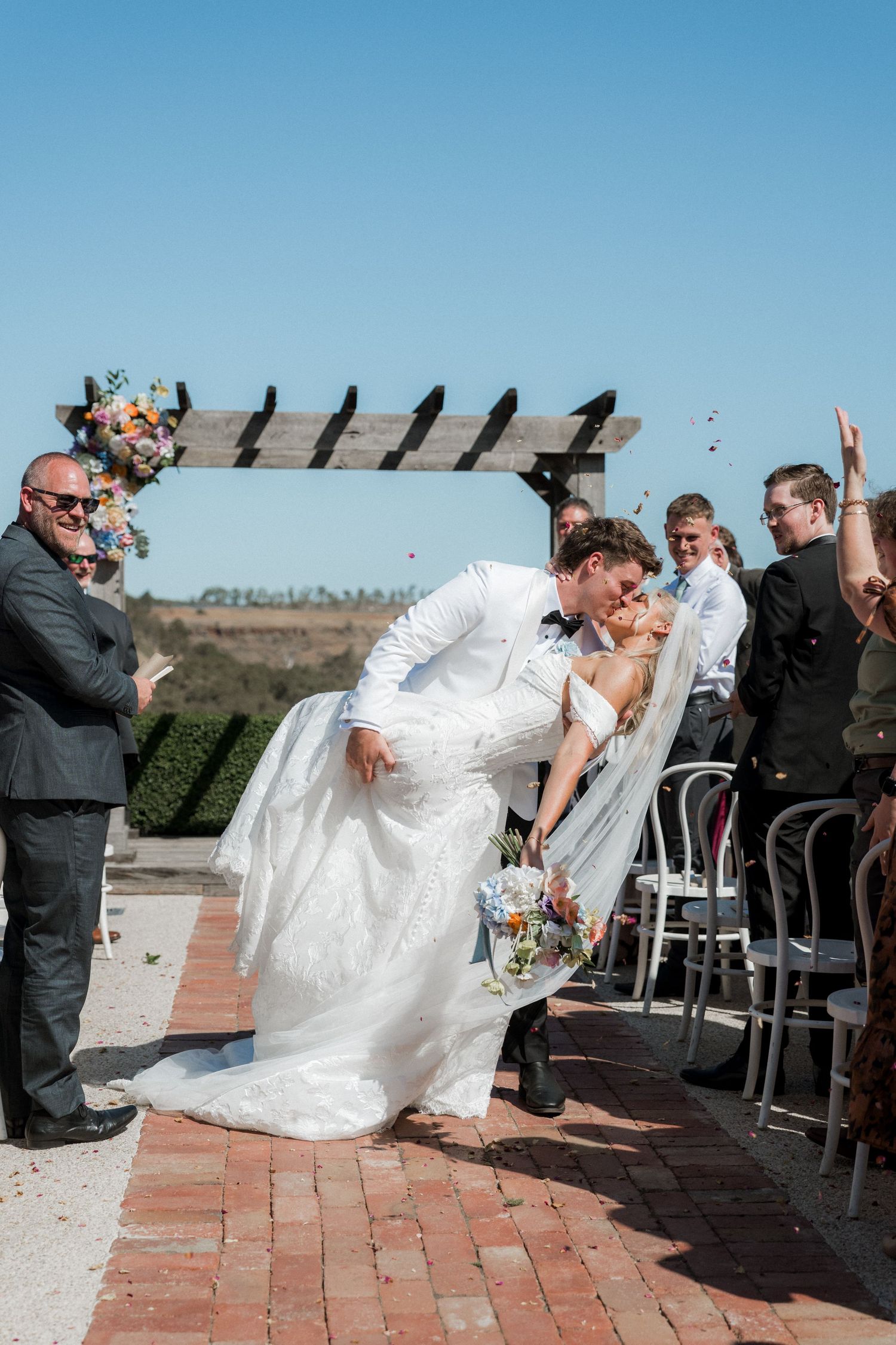 Groom dips bride for a kiss at outdoor wedding ceremony while guests celebrate.