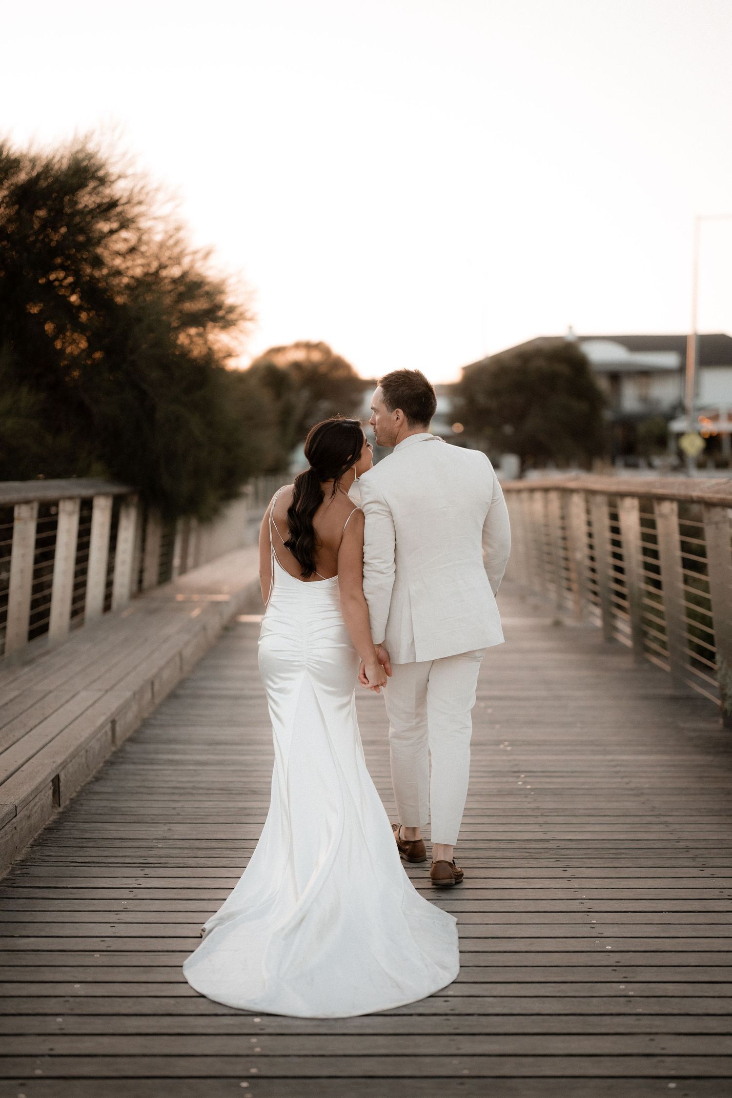 Couple in wedding attire walking together on a wooden boardwalk at sunset.