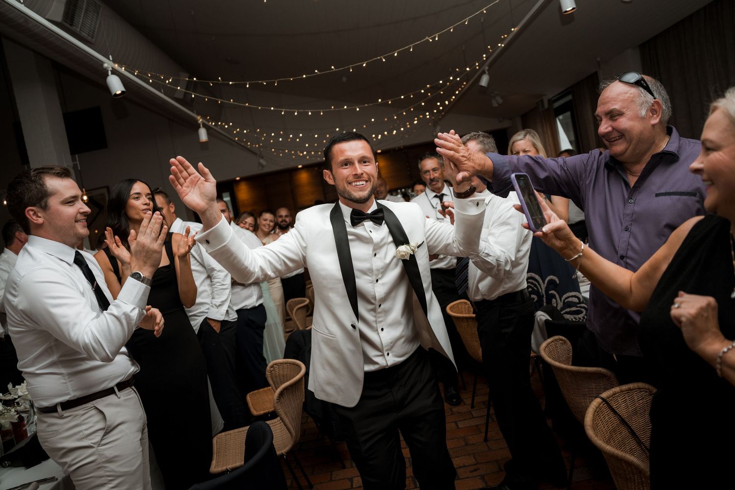 Wedding guests and groomsmen celebrate on the dance floor under twinkling lights at a reception.